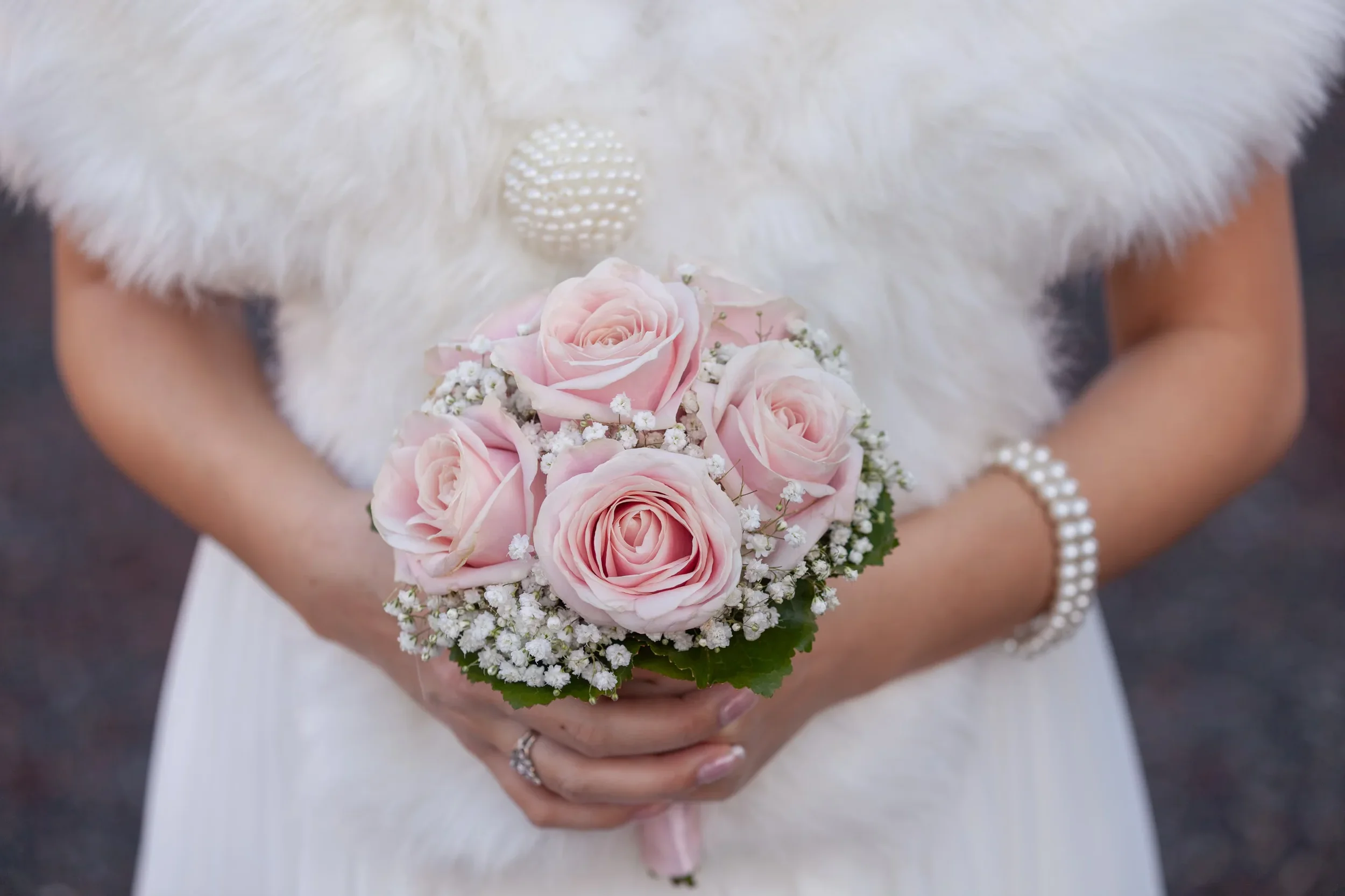 Eine Person hält einen Brautstrauß aus rosa Rosen, weißen Blumen und grünen Blättern. Sie trägt eine weiße Hochzeitskleidung mit einem pelzigen Mantel und eine Perlenarmkette.