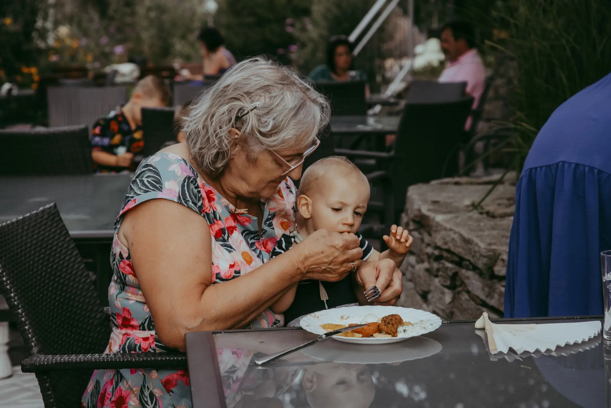 Eine ältere Frau mit grauem Haar und Brille hält ein Kind auf dem Schoß, während sie gemeinsam an einem Essen an einem Tisch im Freien sitzen.