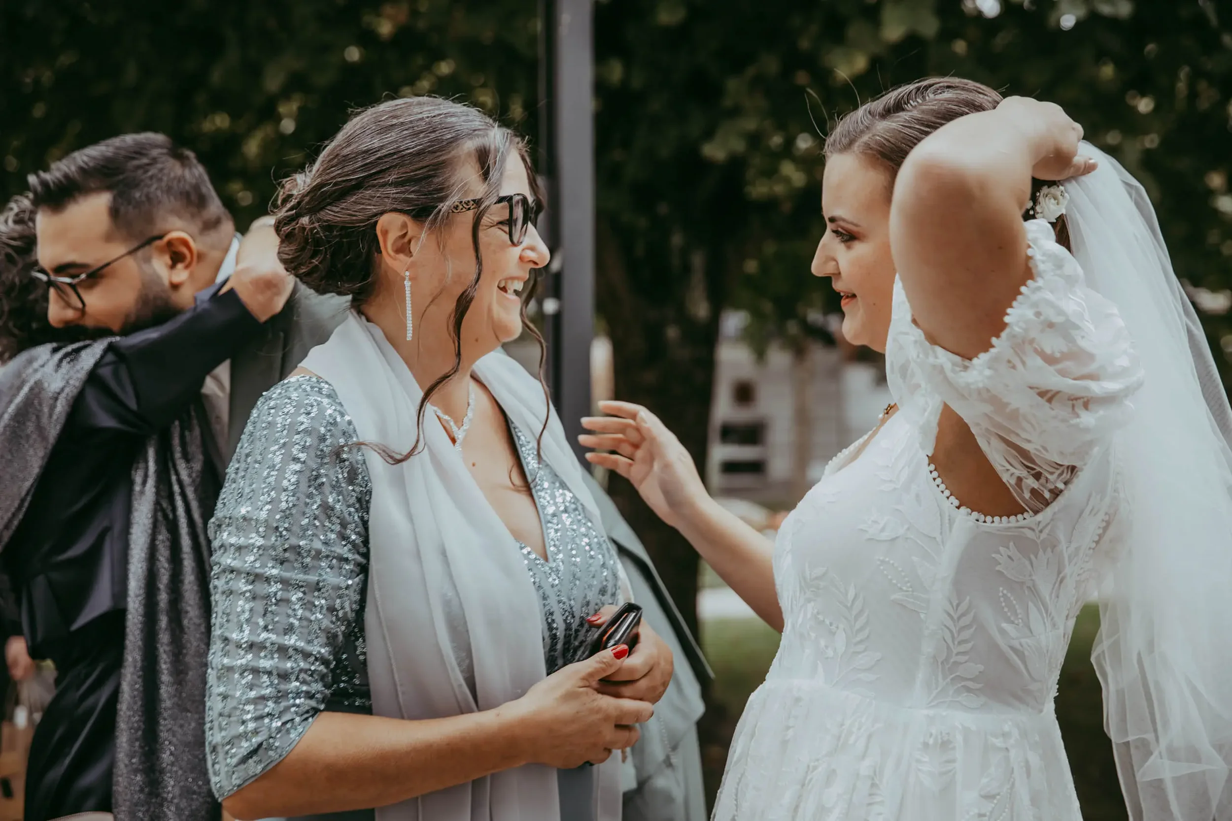 Zwei Frauen bei einer Hochzeit, eine im weißen Brautkleid und die andere in einem silberfarbenen Kleid, lachen sich an, während sie sich begrüßen. Im Hintergrund sind Bäume zu sehen.