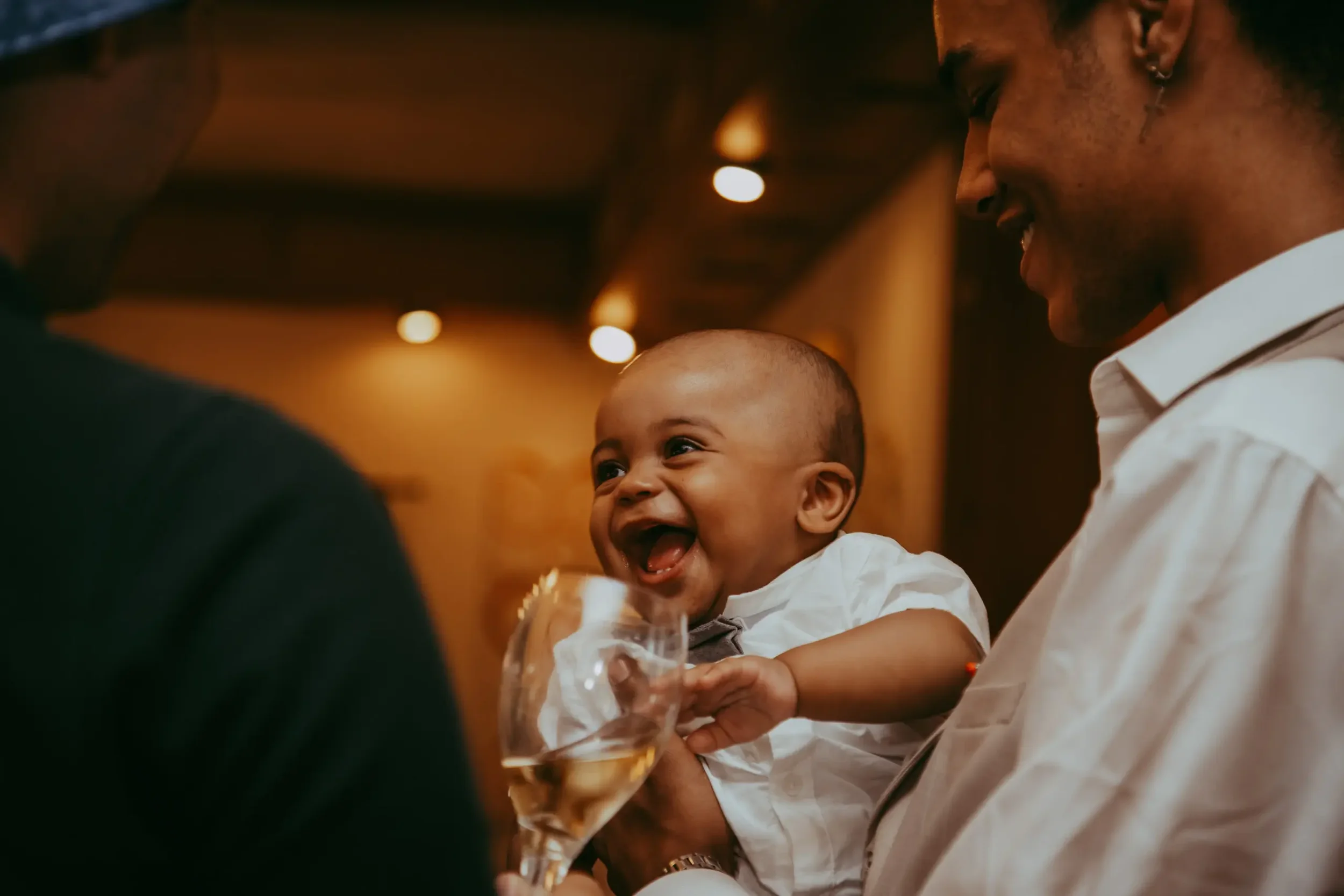 Ein lachendes Baby wird von einer erwachsenen Person gehalten, im Hintergrund eine weitere Person, die einem Glas Champagner hält, in einer warm beleuchteten Umgebung.
