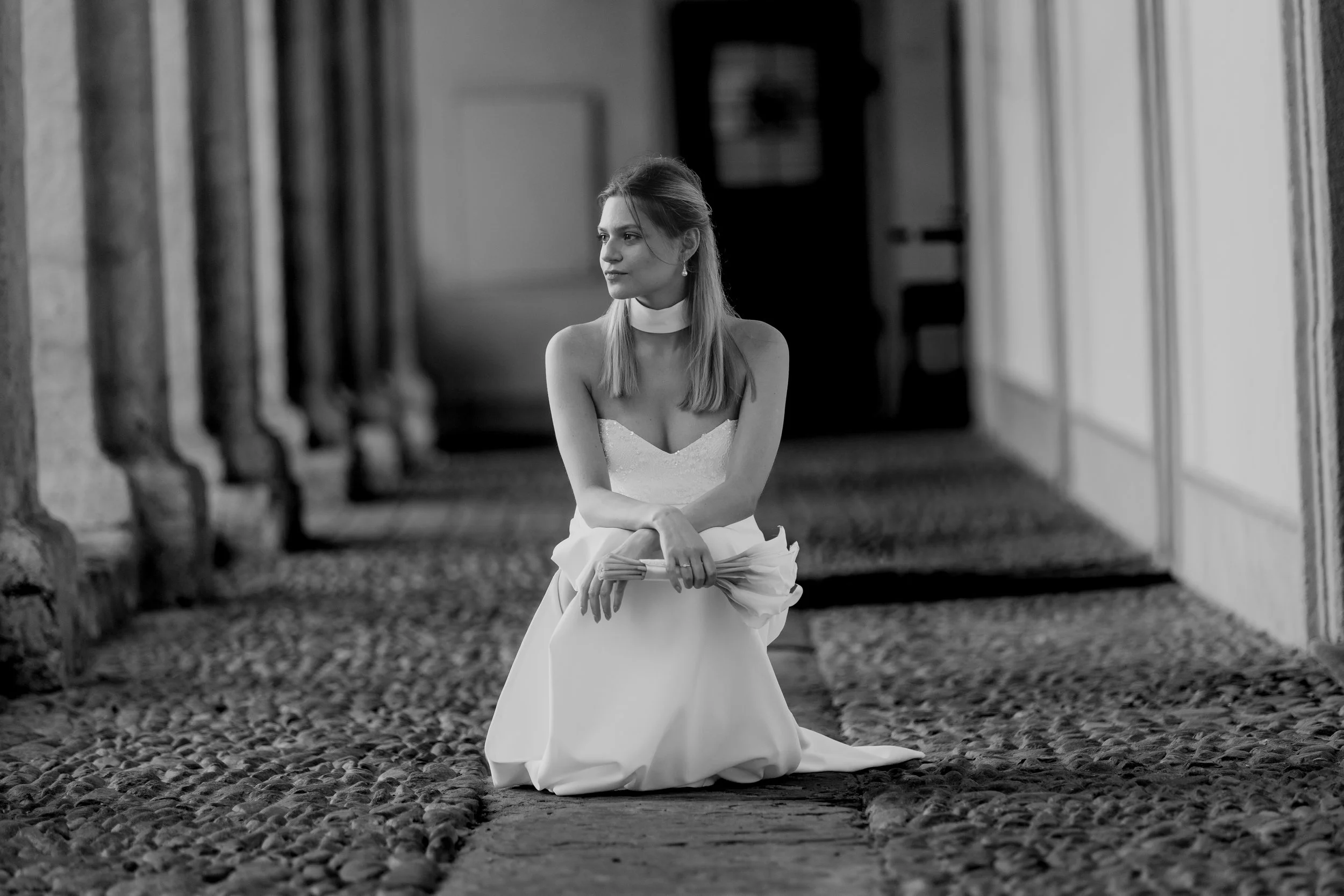Schwarz-weiß-Fotografie einer Frau in einem Brautkleid sitzend auf einem gepflasterten Weg in einem Innenhof, umgeben von Gebäuden mit großen Fenstern und Türen.