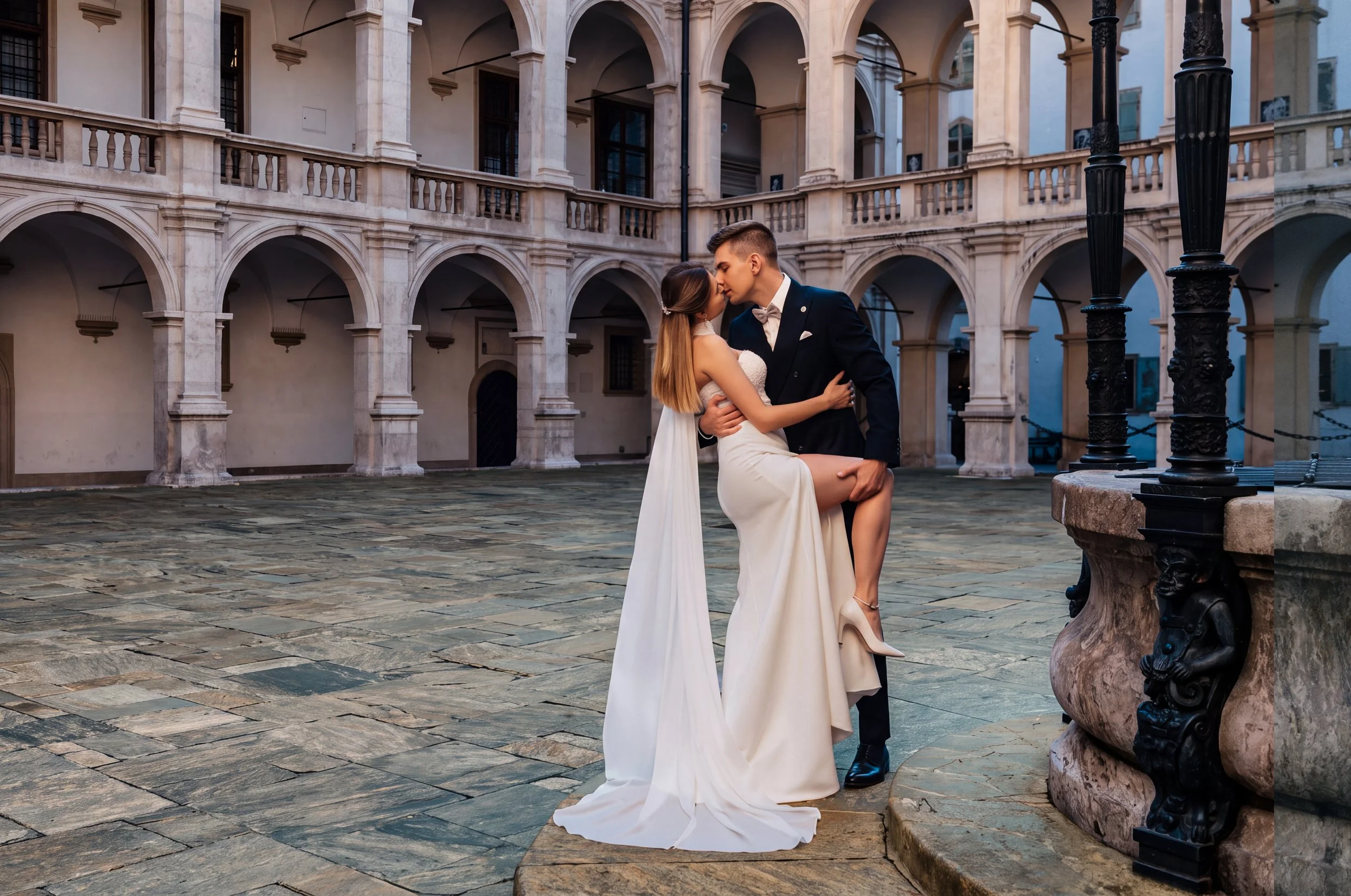 Ein Paar in Hochzeit Kleidung beim Abschiedskuss in einem alten, mehrstöckigen Innenhof mit Bögen und Fenstern. Der Boden ist gepflastert, und es gibt antike Laternen.