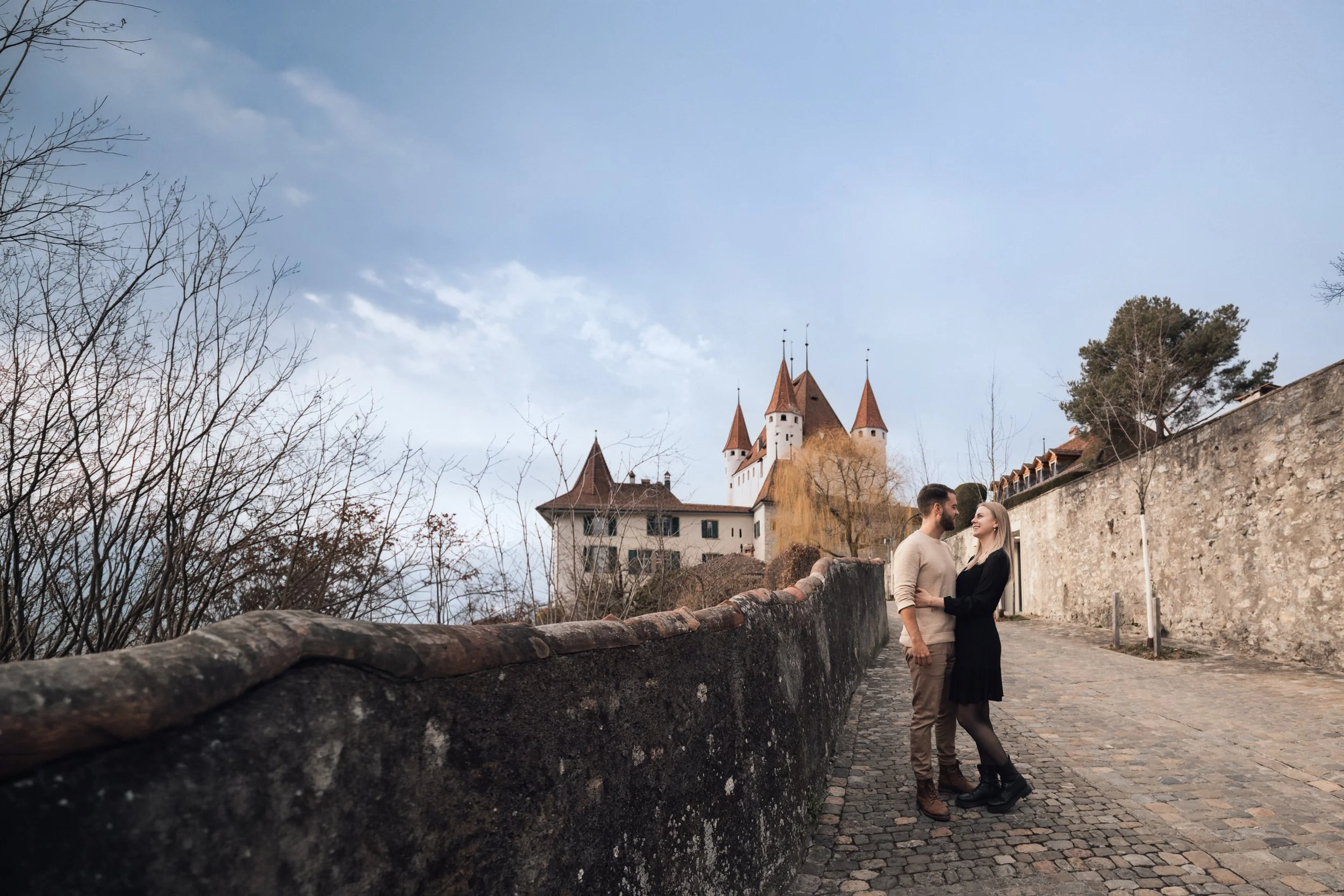 Ein Liebespaar, ein Mann und eine Frau, stehen sich nahe beieinander auf einem gepflasterten Weg vor einer alten Mauer, im Hintergrund eine Burg mit mehreren Türmen und spitzen Dächern, umgeben von Bäumen ohne Blätter sowie einigen hohen Bäumen.