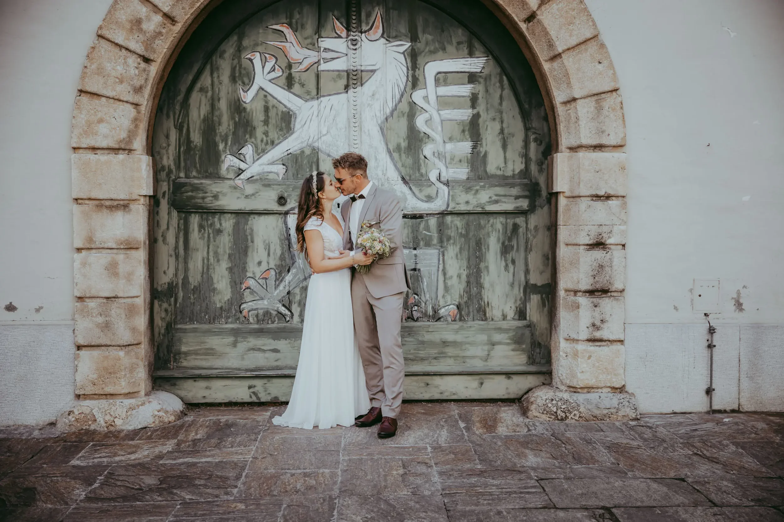 Ein Brautpaar steht vor einer alten Holz- und Stein-Tür mit einer Wandmalerei im Hintergrund. Die Frau trägt ein langes weißes Hochzeitskleid und hält einen Blumenstrauß, der Mann trägt einen grau Anzug mit Fliege.