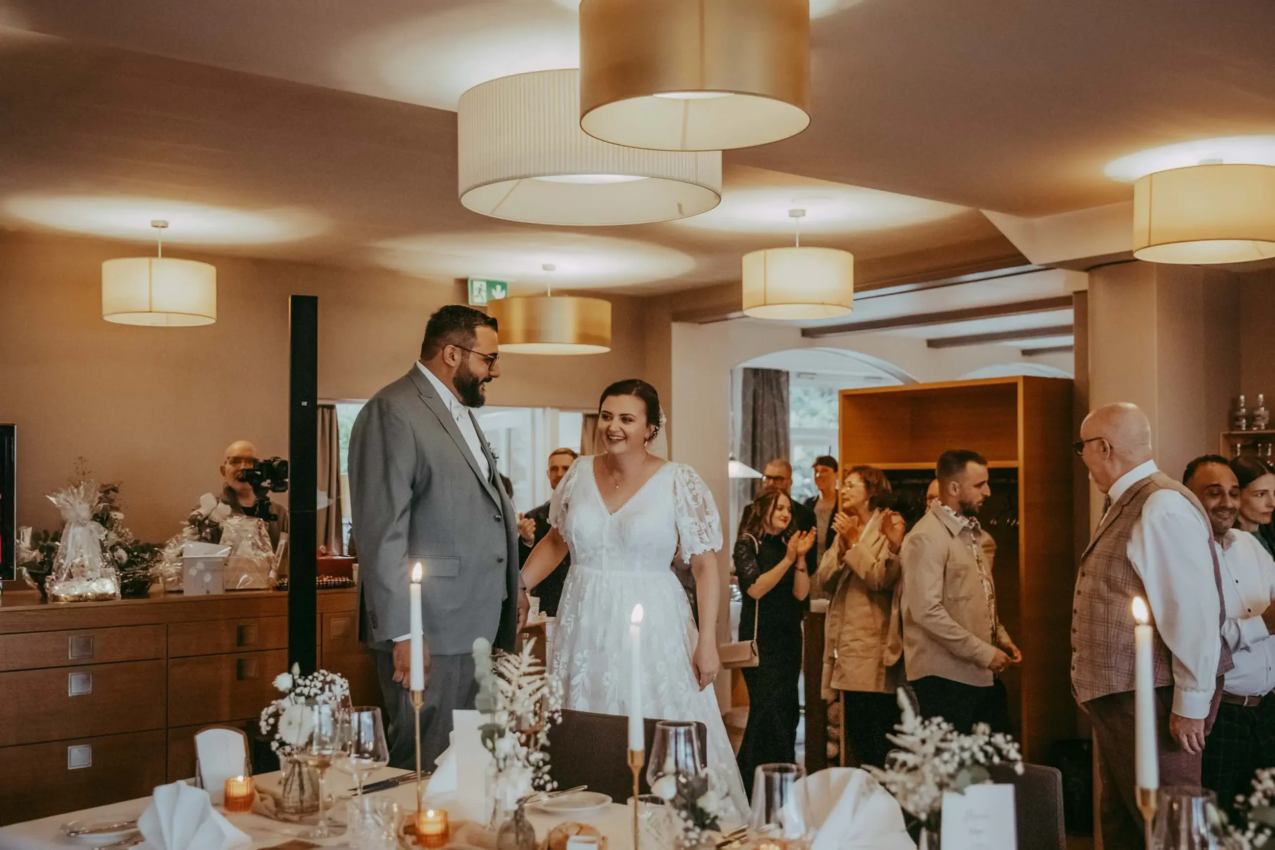 Ein junges Paar bei einer Hochzeit, umgeben von Gästen in einem festlich dekorierten Raum mit Kerzen, lieblichem Licht und Tischen.