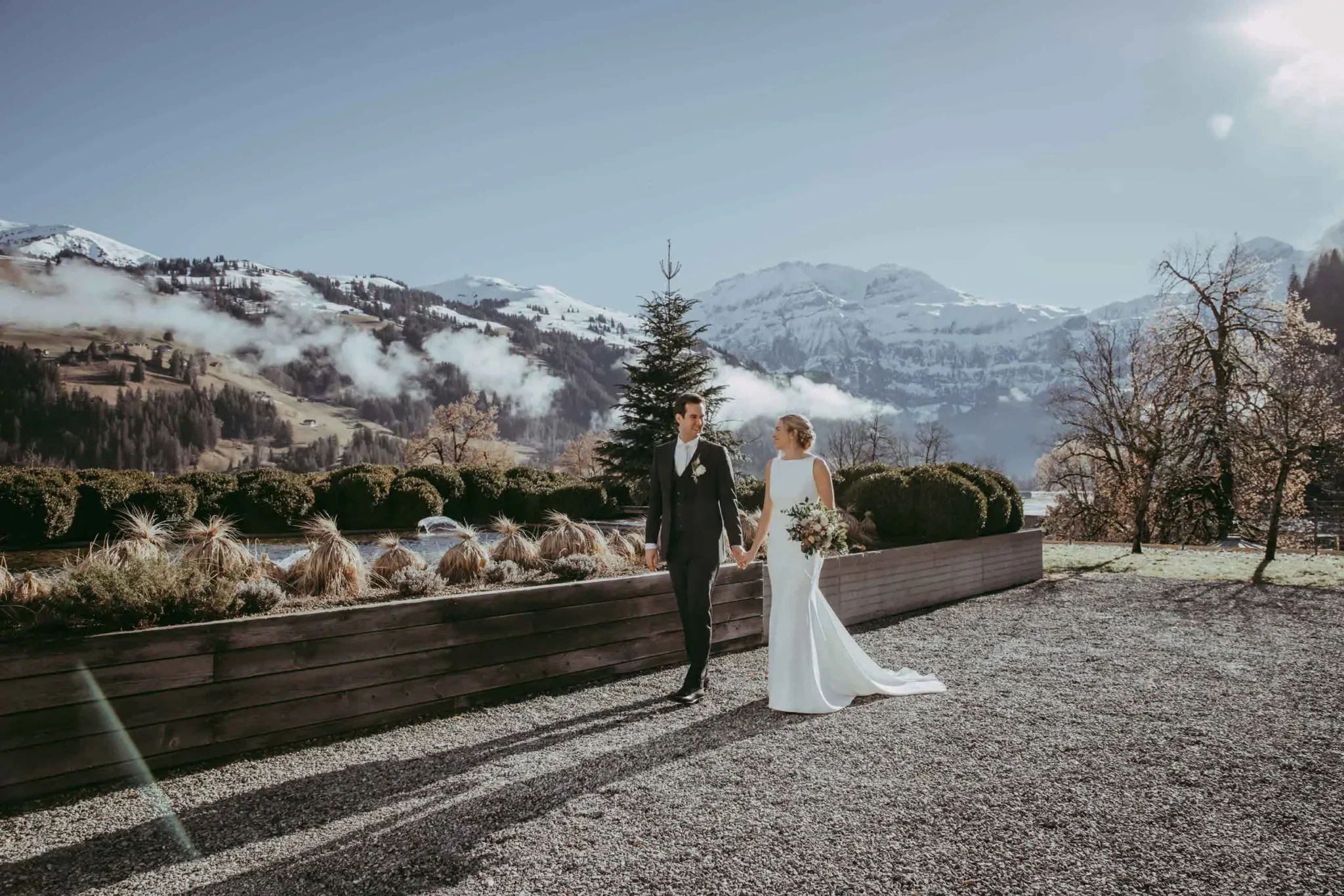 Ein Brautpaar beim Spaziergang am Tag ihrer Hochzeit mit Bergen im Hintergrund, sonnigem Himmel und Bäumen.