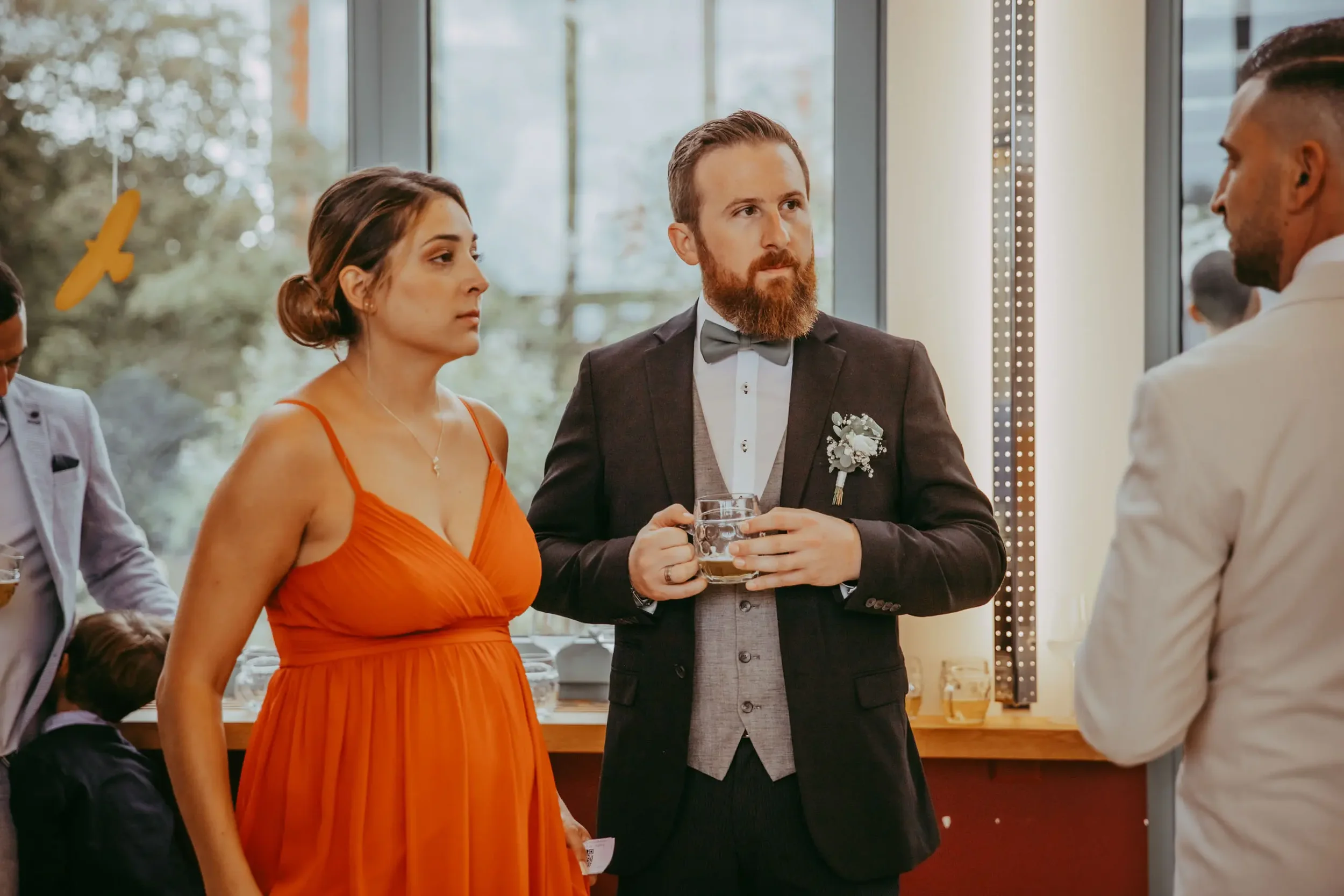 Menschen bei einer formellen Veranstaltung im Raum, eine Frau in einem orangefarbenen Kleid und ein Mann mit Anzug und Krawatte halten Gläser, während sie sprechen.