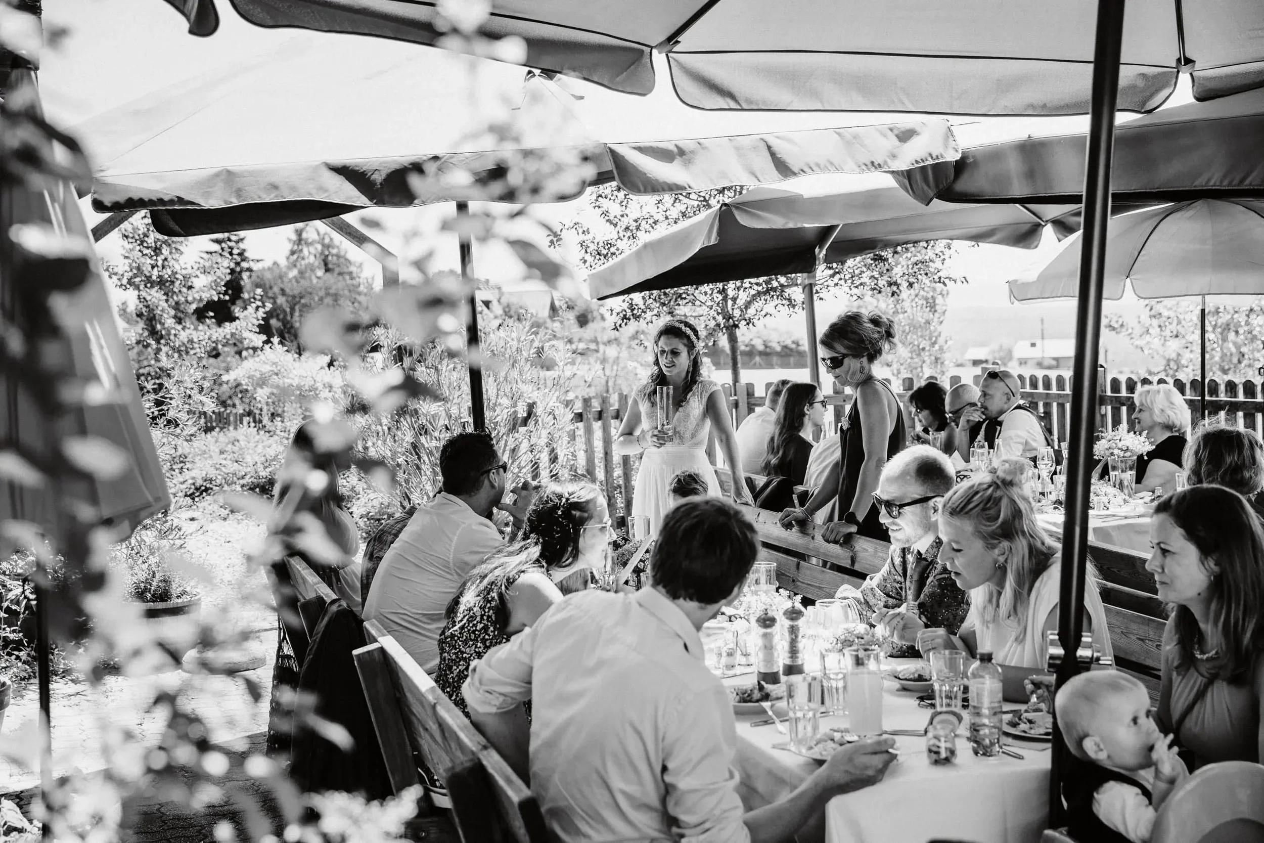 Menschen sitzen an einem langen Gartentisch im Freien, essen und trinken, während im Hintergrund eine Frau in einem Kleid steht und spricht.