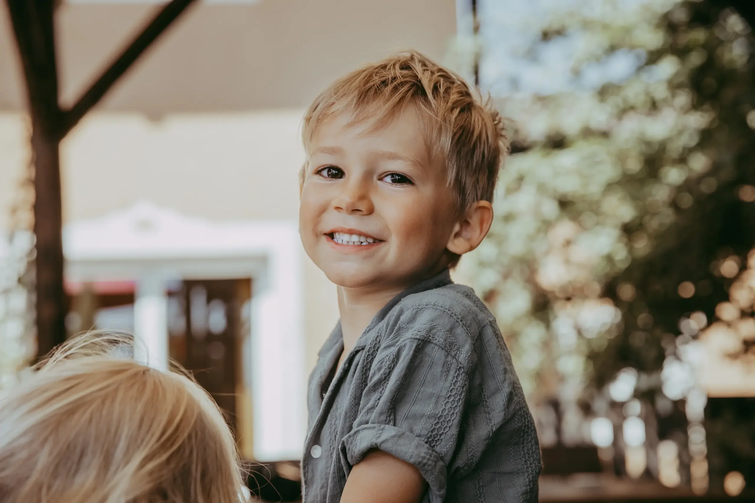 Ein jüngerer Jungen mit blonden Haaren, der draußen vor buntem Hintergrund mit Bäumen steht und lächelt.