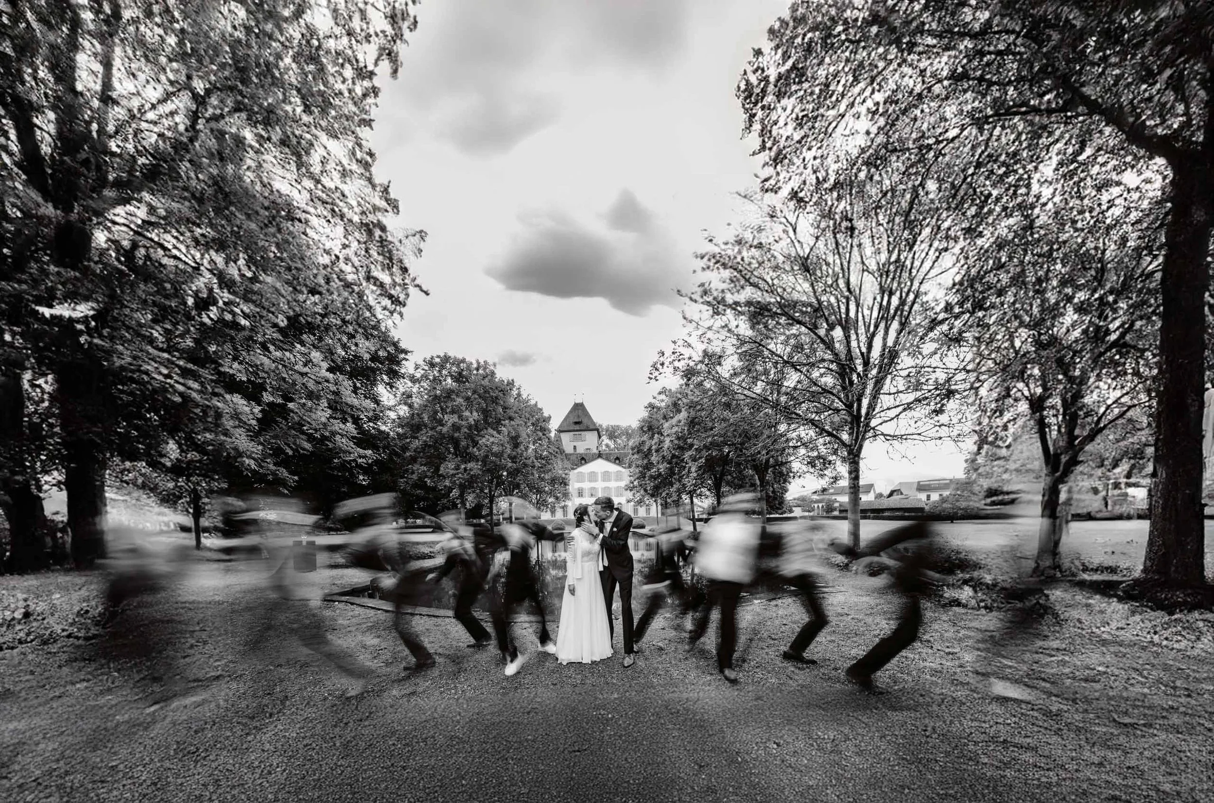 Schwarz-weiß-Foto eines Paares, das sich in einem Park bei Regen umarmt, während sie Regenschirme tragen. Im Hintergrund Bäume und ein Gebäude mit Turm. Die Szene ist romantisch und idyllisch.