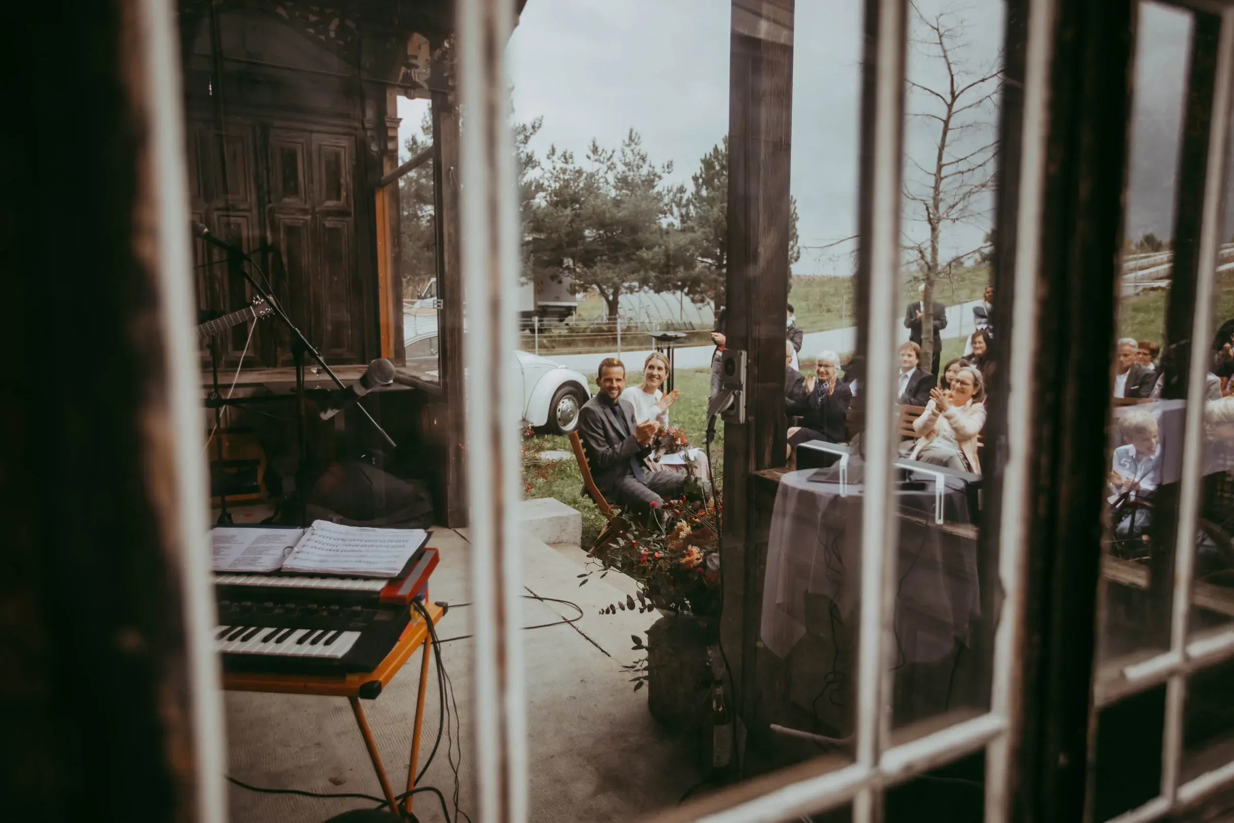 Menschen sitzen bei einer Outdoor-Veranstaltung, gesehen durch eine Gittertür, bei der es aussieht wie eine Musik- oder Feierveranstaltung, mit einem Klavier im Vordergrund.