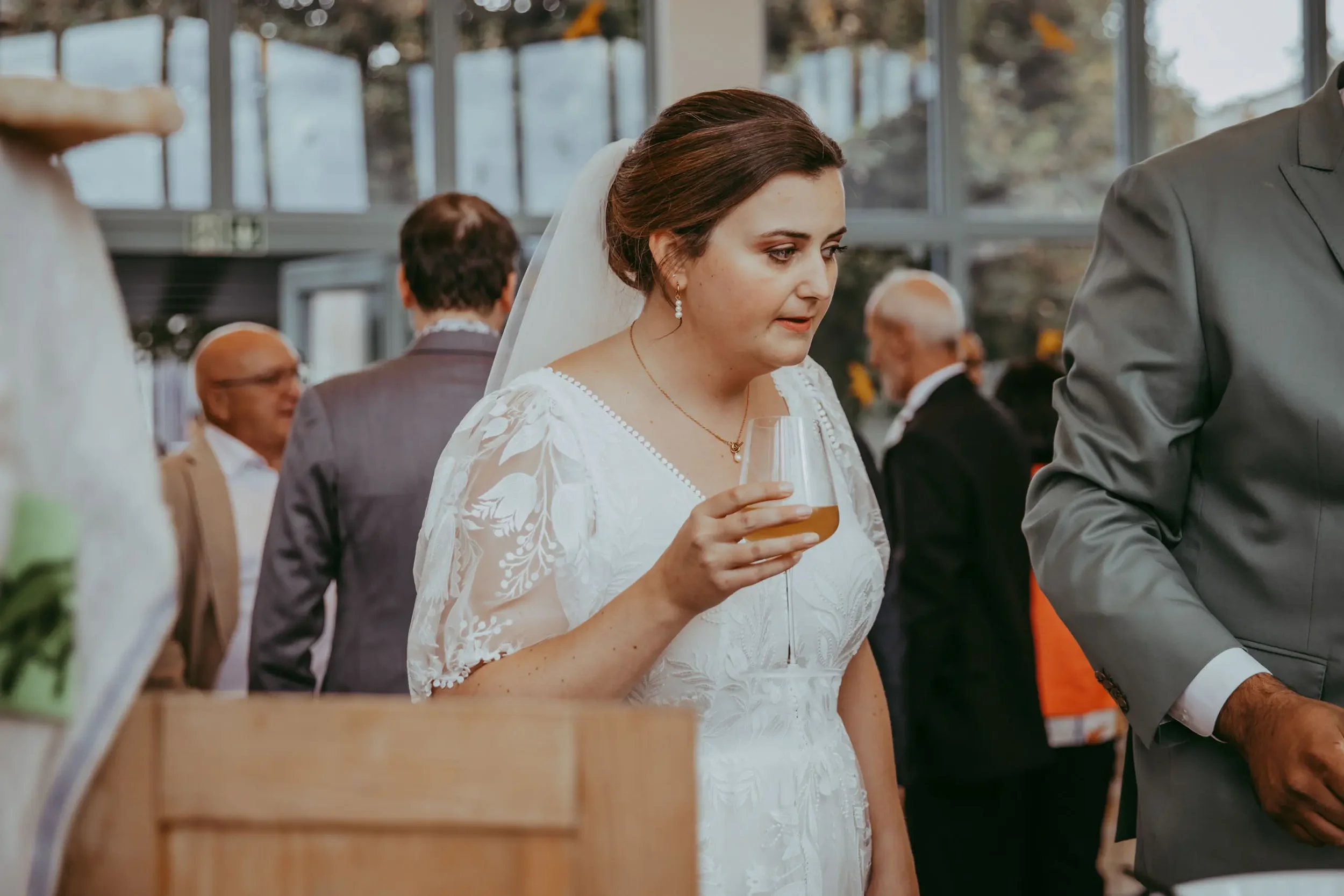 Eine Braut in einem weißen Kleid hält ein Weinglas und steht in einer Hochzeitshalle, umgeben von Gästen, die sich unterhalten.
