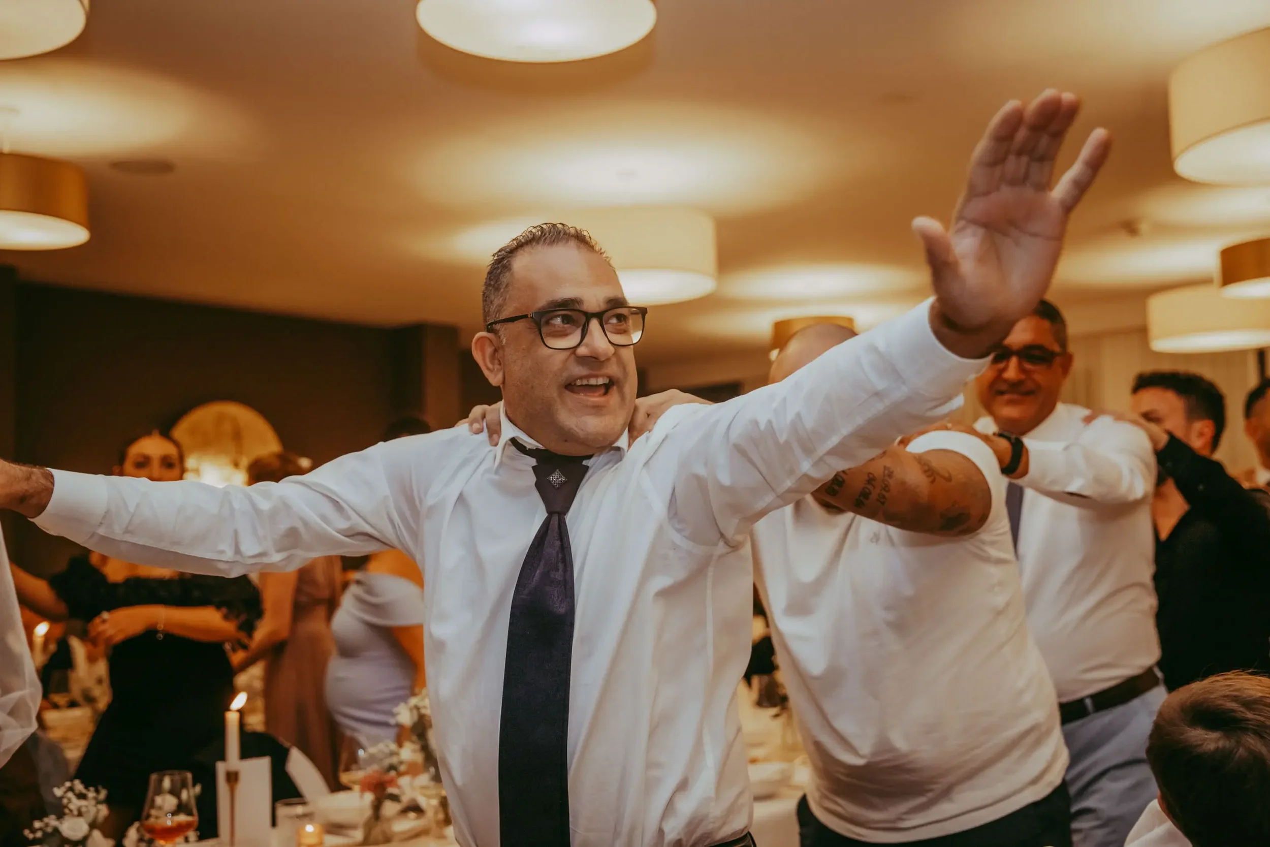 Ein Mann mit Brille und Krawatte tanzt auf einer Party, Menschen um ihn herum feiern in einem warm beleuchteten Raum.
