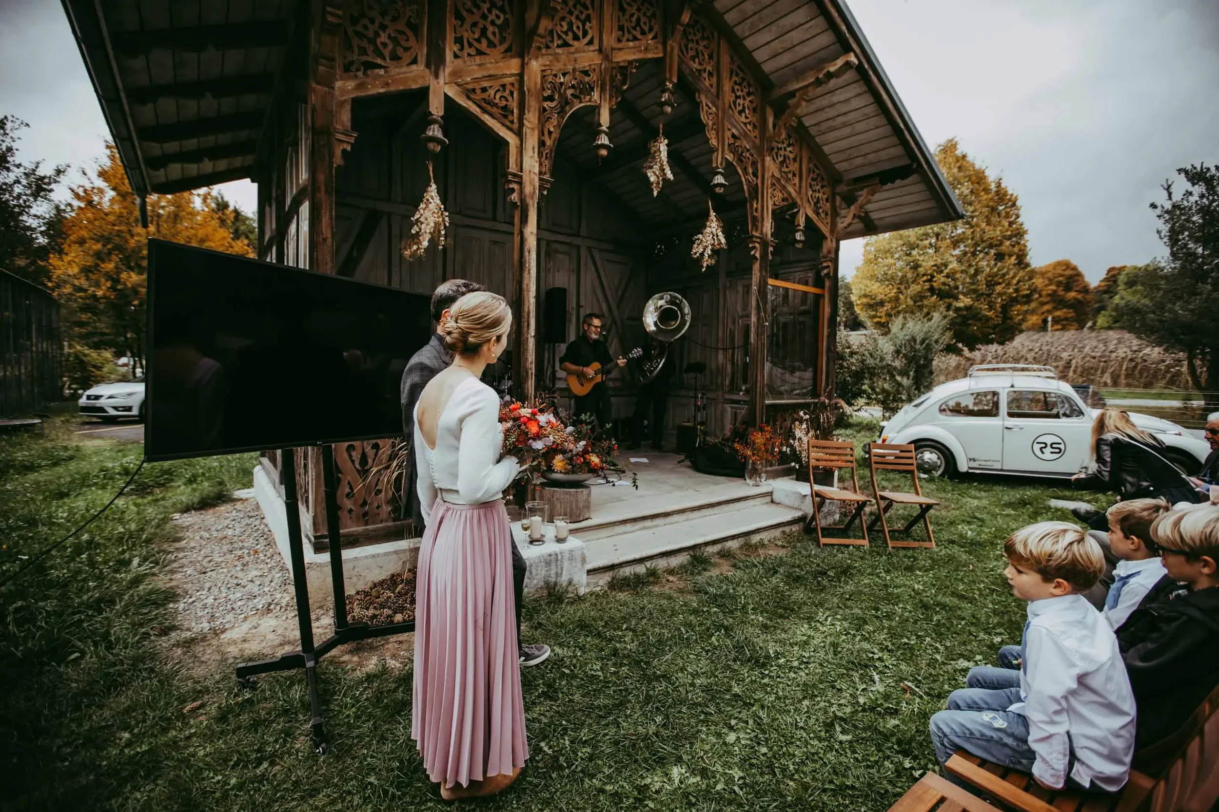 Festliche Musiker spielen auf einer kleinen Bühne im Freien bei einer Hochzeit, im Hintergrund bunte Herbstbäume.