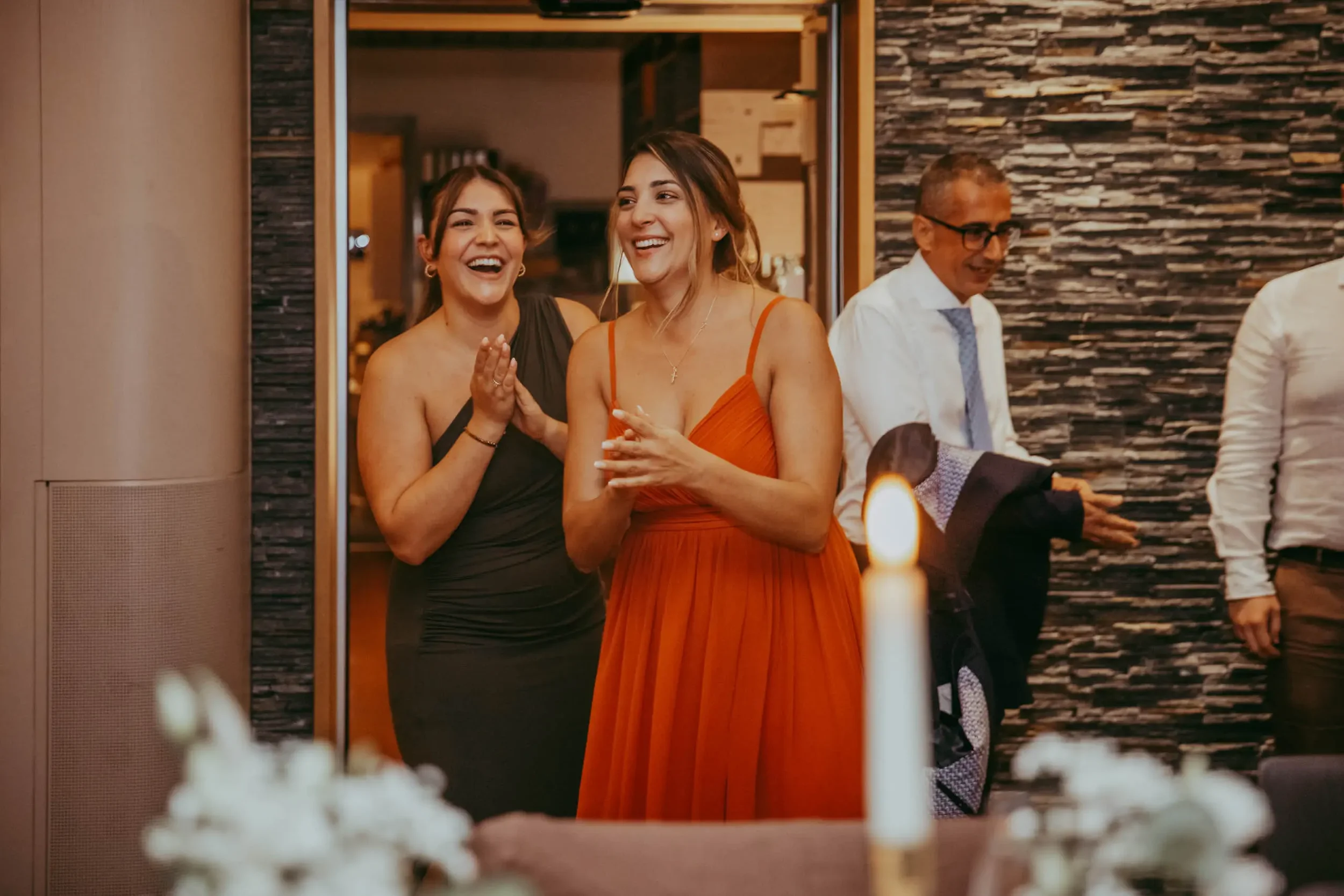Zwei lachende Frauen in eleganten Kleidern feiern in einem festlichen Raum, während ein älterer Herr im Hintergrund steht.