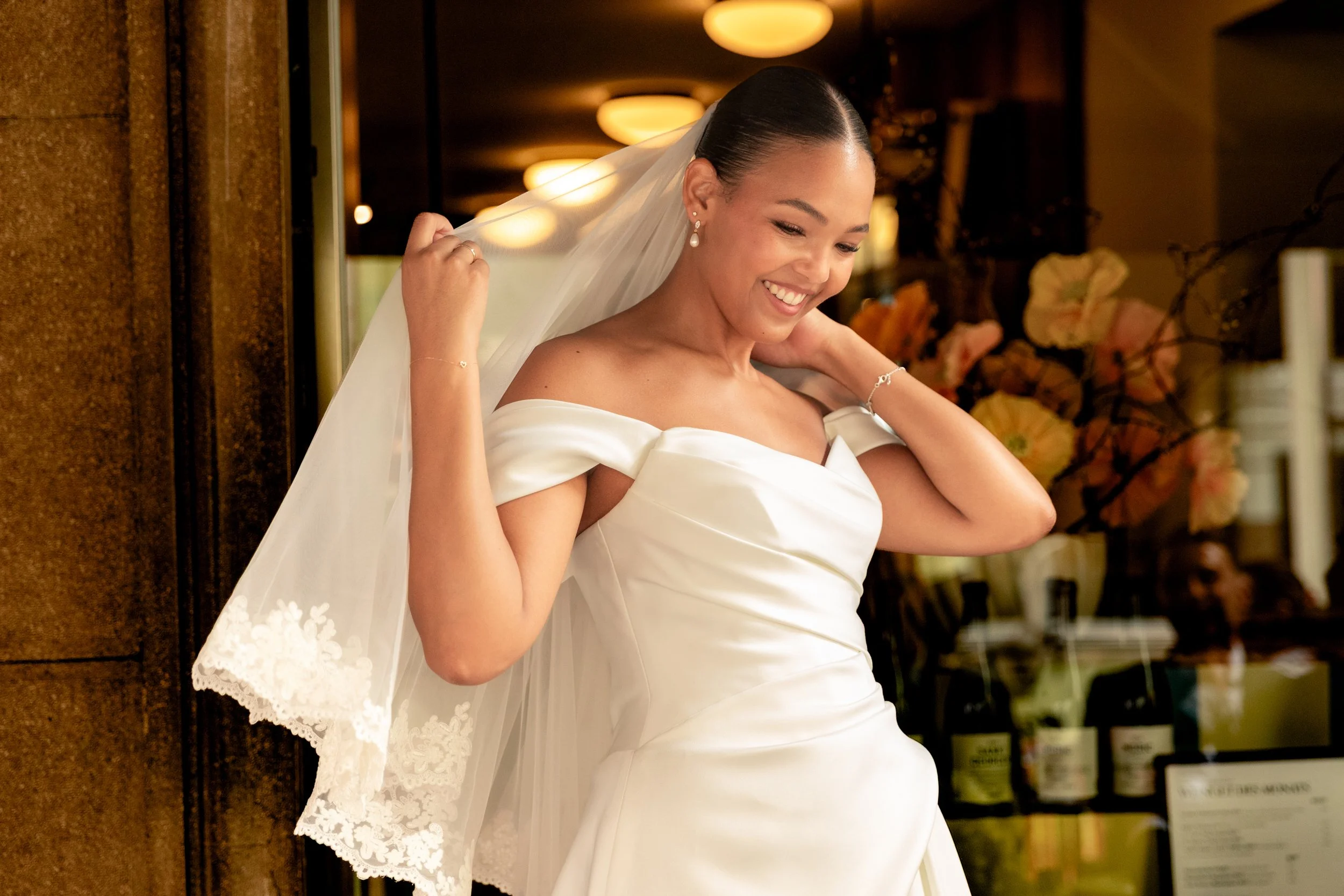 Eine lachende Frau in einem weißen Hochzeitskleid mit Schleier und Perlenohrringen, die an einer Tür steht, umgeben von einem warm beleuchteten Raum.