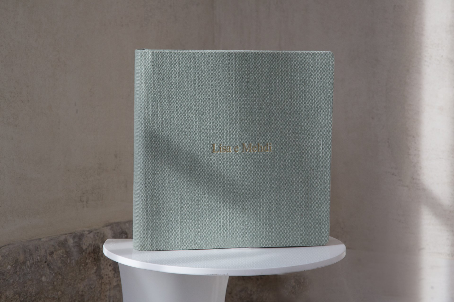 Ein graues, stoffbezogenes Buch mit goldener Schrift, auf einem weißen Tisch gegen eine graue Wand. Der Titel lautet 'Lisa e Mehdi'.