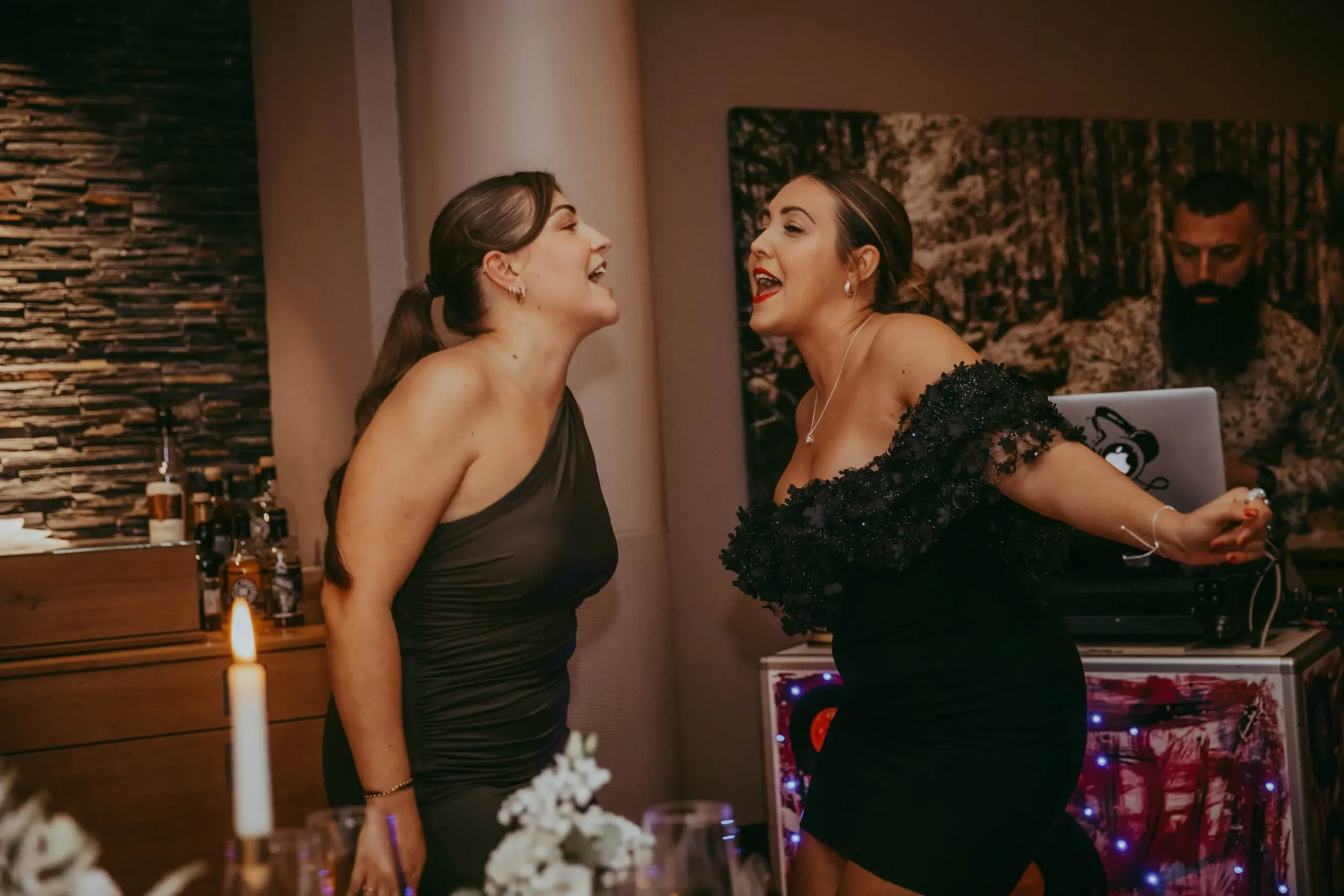 Zwei Frauen, die auf einer Party lachen und sich nahe kommen, während ein DJ im Hintergrund Musik spielt.