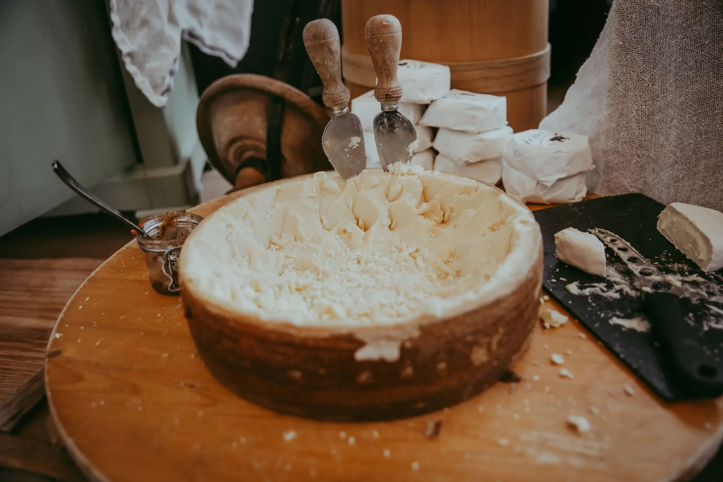 Haufen von Käse in einer Holschale, zwei Käsemesser stecken im Käse, umgeben von Käseblöcken und -scheiben, auf einem Holzbrett.