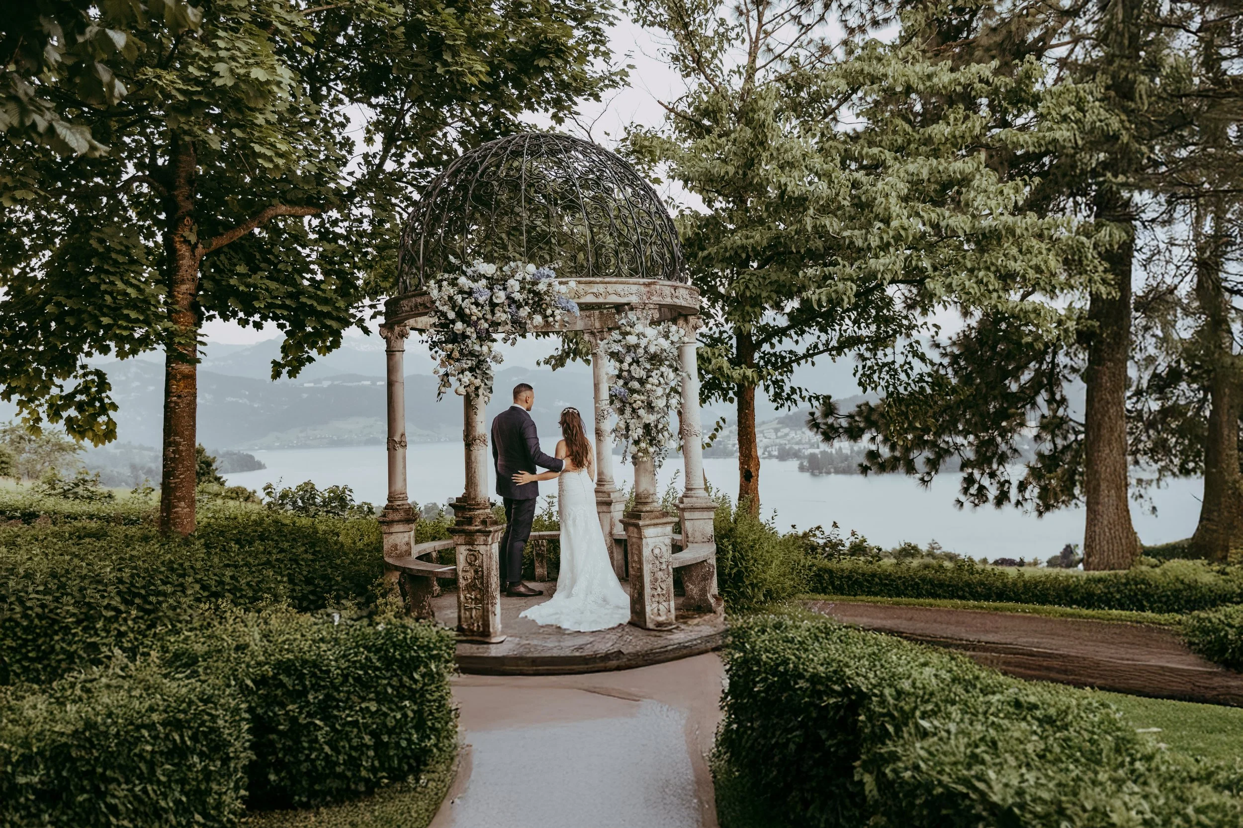 Ein Brautpaar steht unter einem kleinen Pavillon mit verzierter Metallkonstruktion, umgeben von Bäumen, bei einer Hochzeit an einem See.