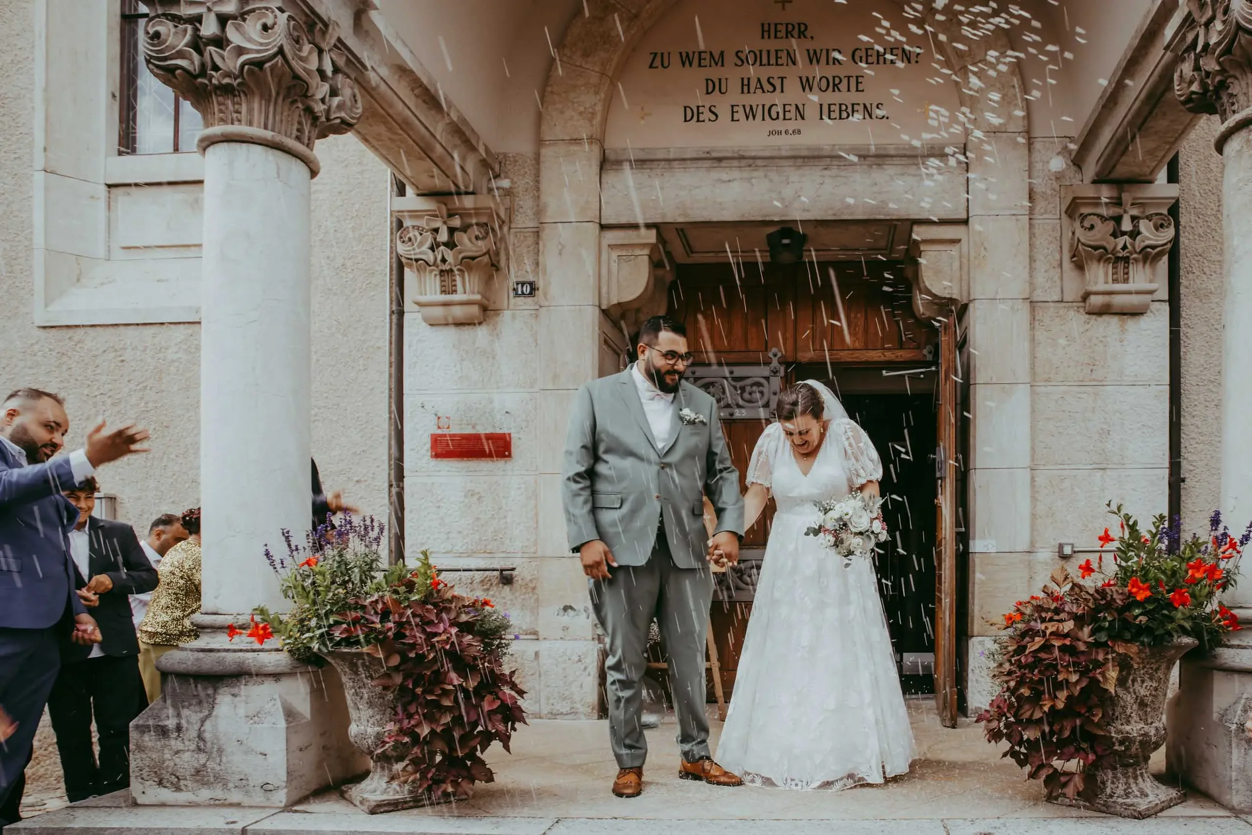 Ein Brautpaar und Gäste vor einer Kirche, bei einer Hochzeit, während des Regens, mit Text auf der Kirche, die auf Deutsch ist.