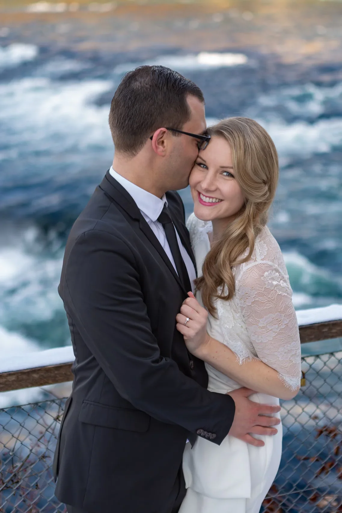 Ein glückliches Paar in Hochzeitskleidung steht eng aneinandergeschlungen am Meer, mit Wasser im Hintergrund, bei Sonnenuntergang.