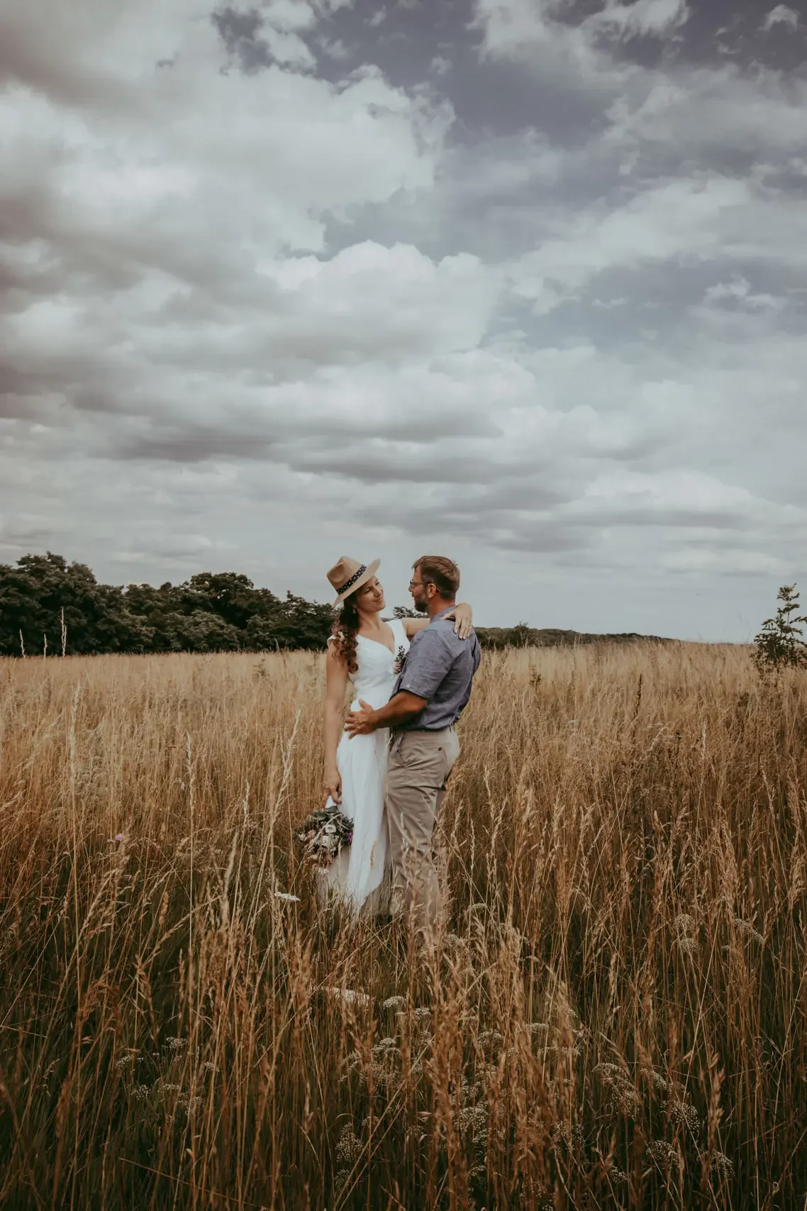 Ein heiratswilliges Paar steht in einem trockenen Feld unter einem bewölkten Himmel und schaut sich verliebt an.