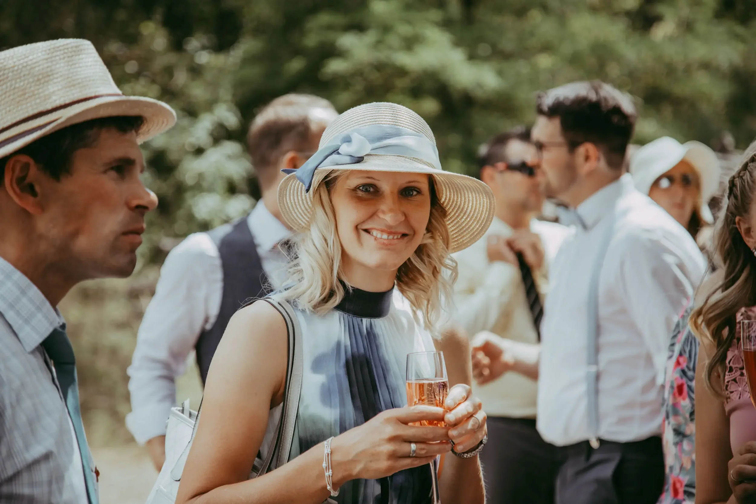 Eine Gruppe von Menschen bei einer outdoor Veranstaltung, eine lachende Frau trägt einen Hut mit Schleife und hält ein Glas Sekt.