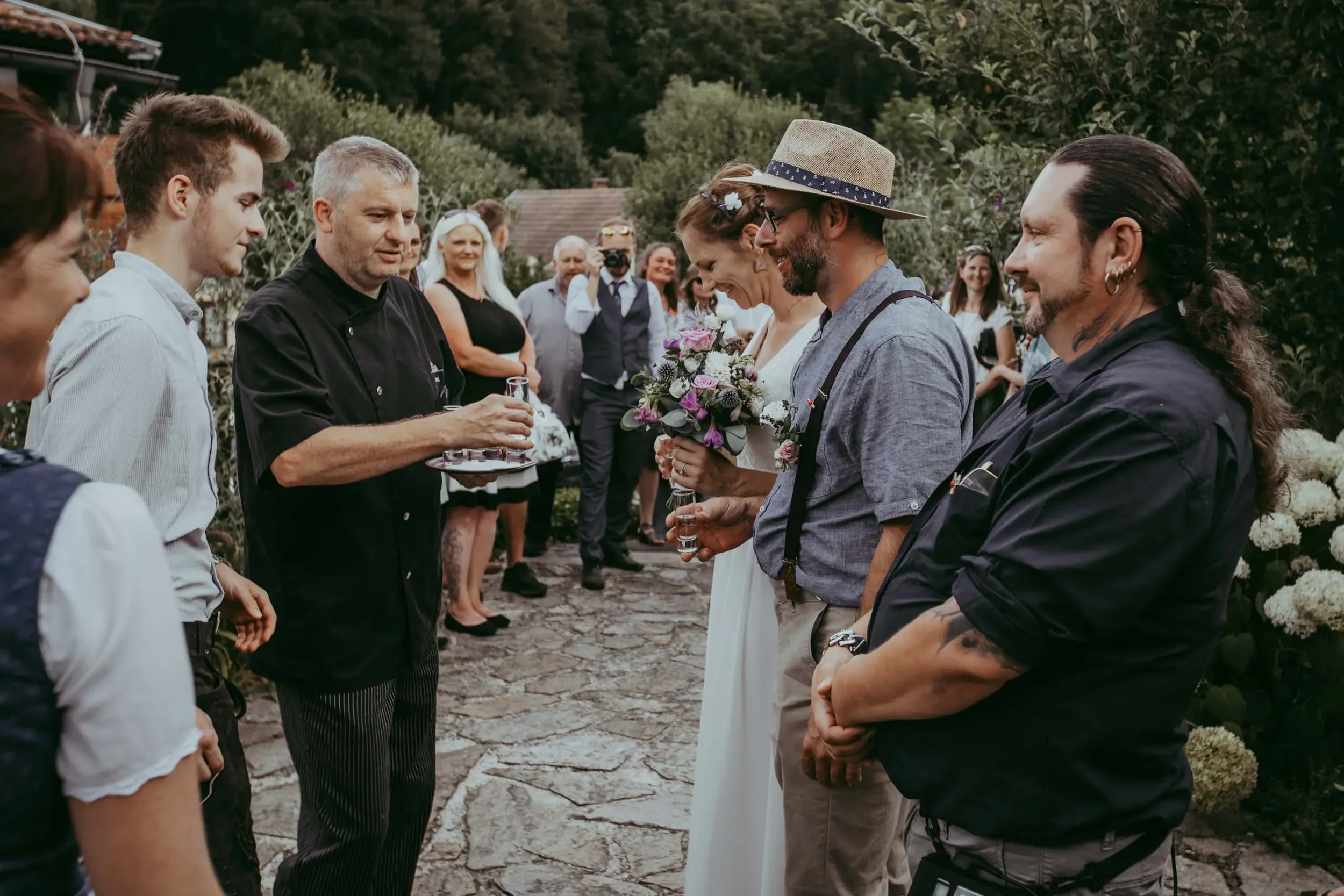Hochzeit im Freien mit einer Gruppe von Menschen, die sich um die Braut und den Bräutigam versammelt haben, mit Blumen und Natur im Hintergrund.