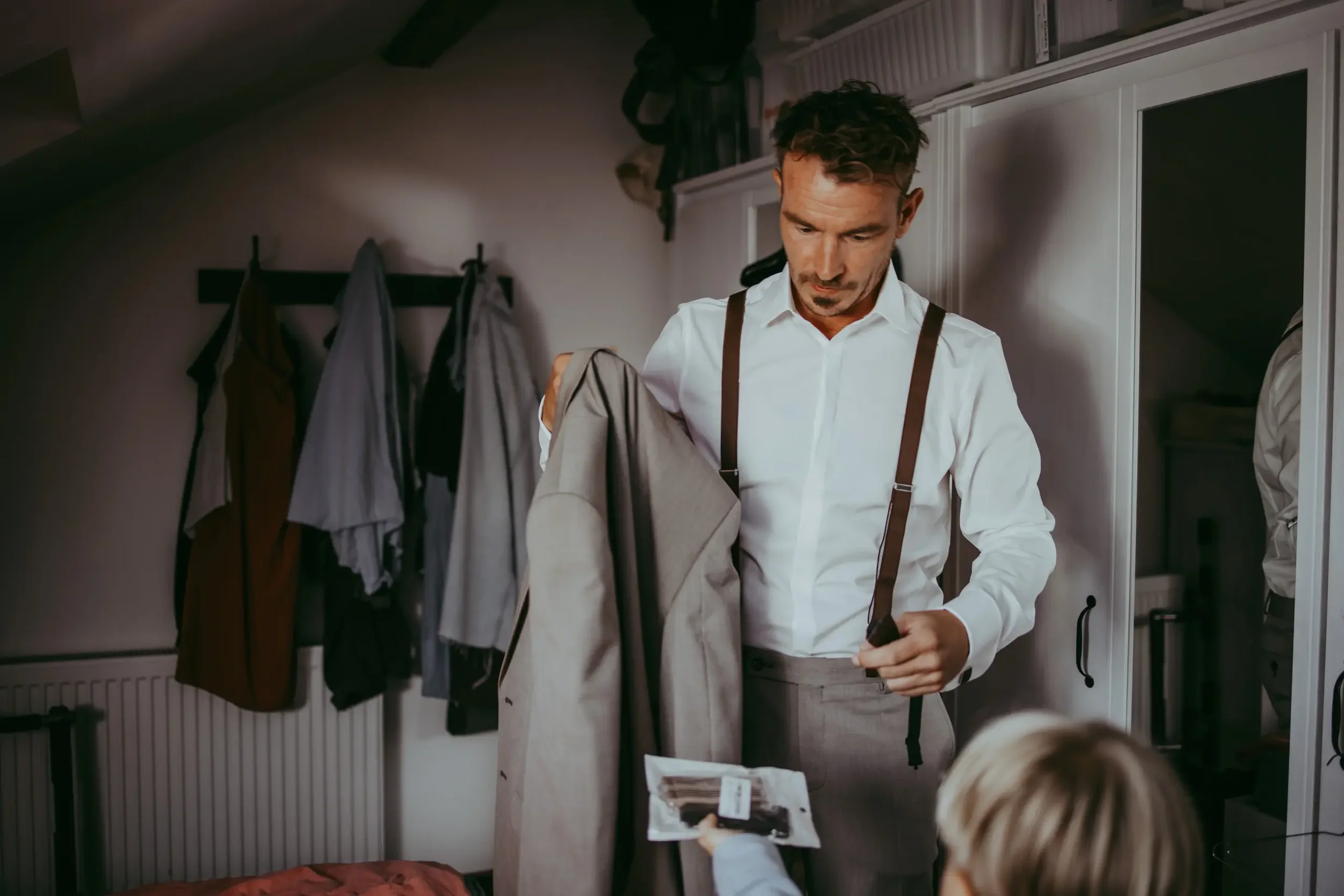 Ein Mann in weißen Hemd und Hosenträger richtet Kleidung im Schlafzimmer. Im Vordergrund hält ein Kind mit langem, blondem Haar eine Zeitschrift. Im Hintergrund ist eine Garderobe mit Mänteln und Handtüchern zu sehen.