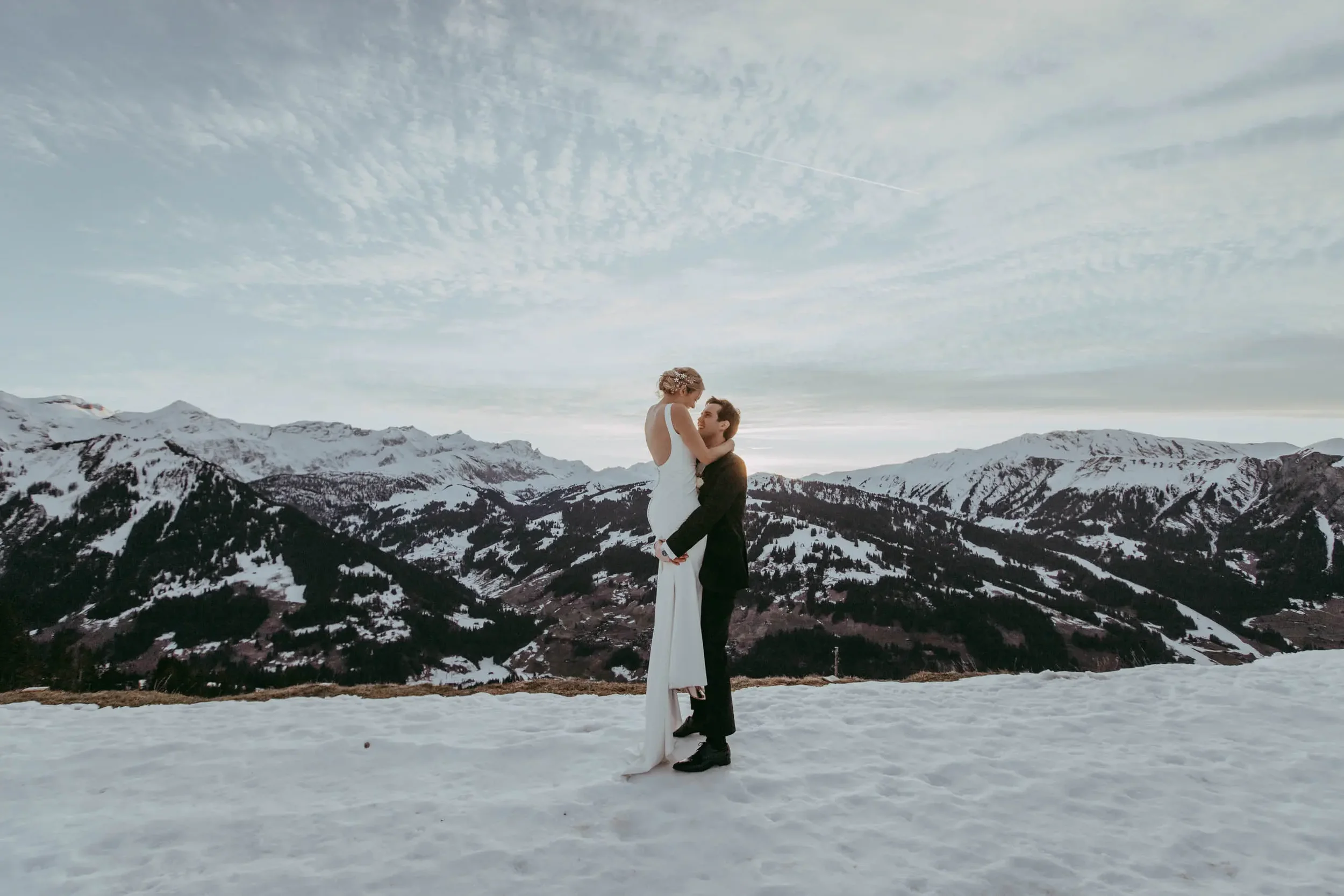 Ein Paar beim Hochzeitsfoto in schneebedeckten Bergen, die Frau trägt ein weißes Brautkleid und der Mann einen Anzug, sie umarmen sich vor der winterlichen Landschaft.