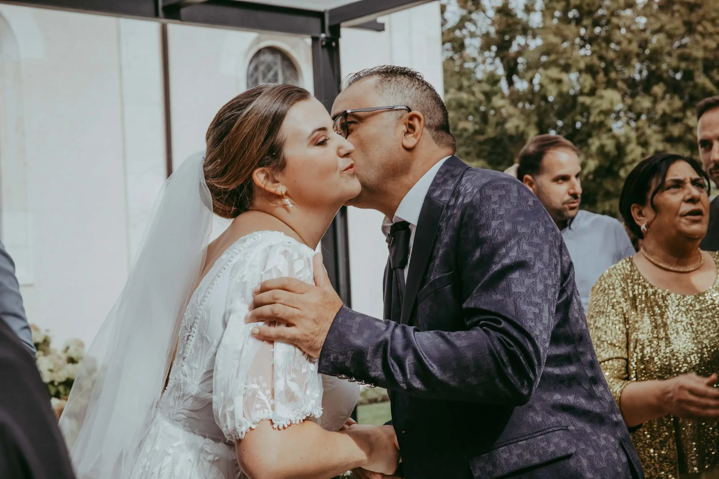 Ein Brautpaar bei einer Hochzeit, küsst sich im Freien, umgeben von Gästen und Bäumen.