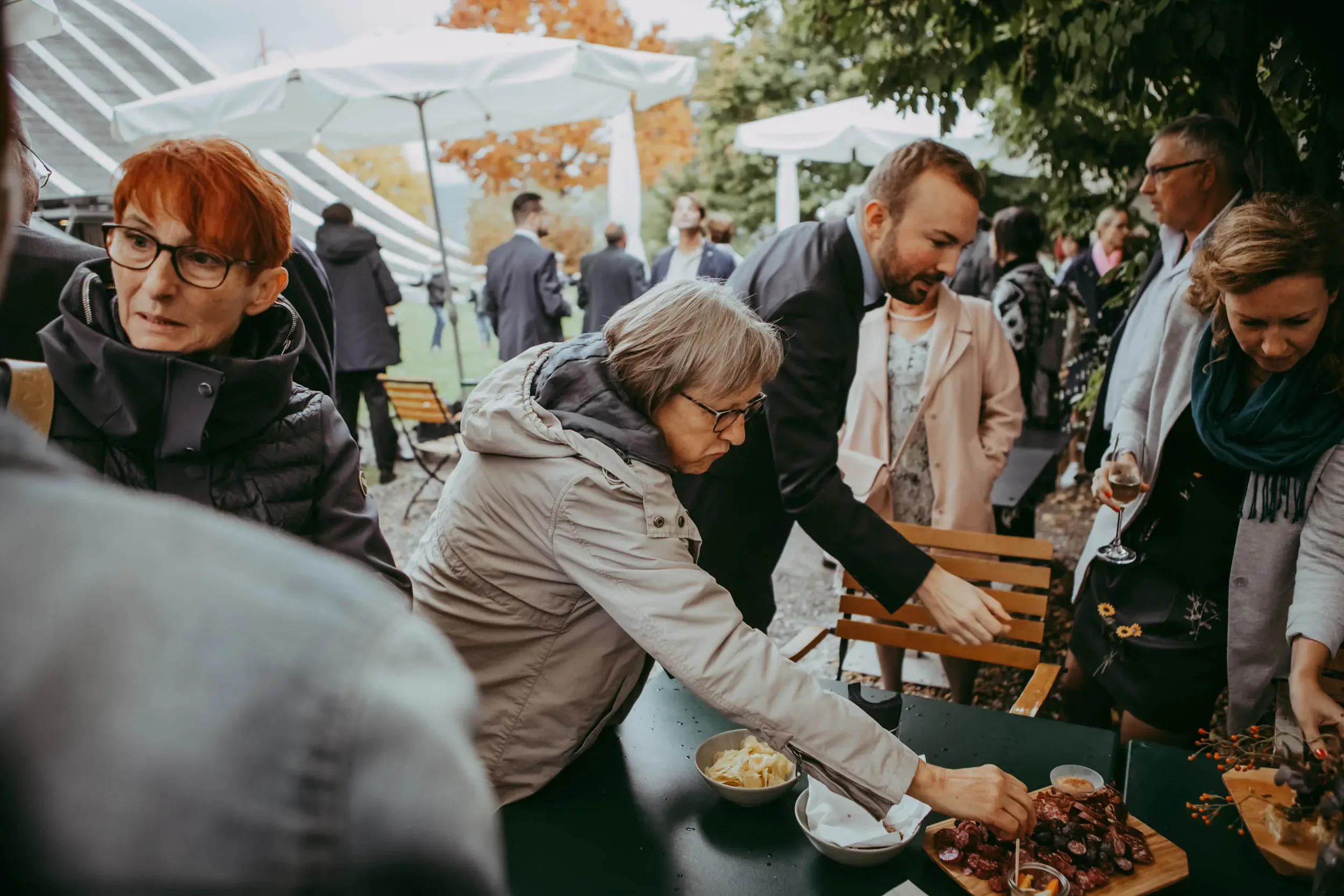 Menschen bei einem Outdoor-Empfang, die sich um eine Tafel mit Snacks und Getränken versammeln, unter Bäumen bei sonnigem Wetter.