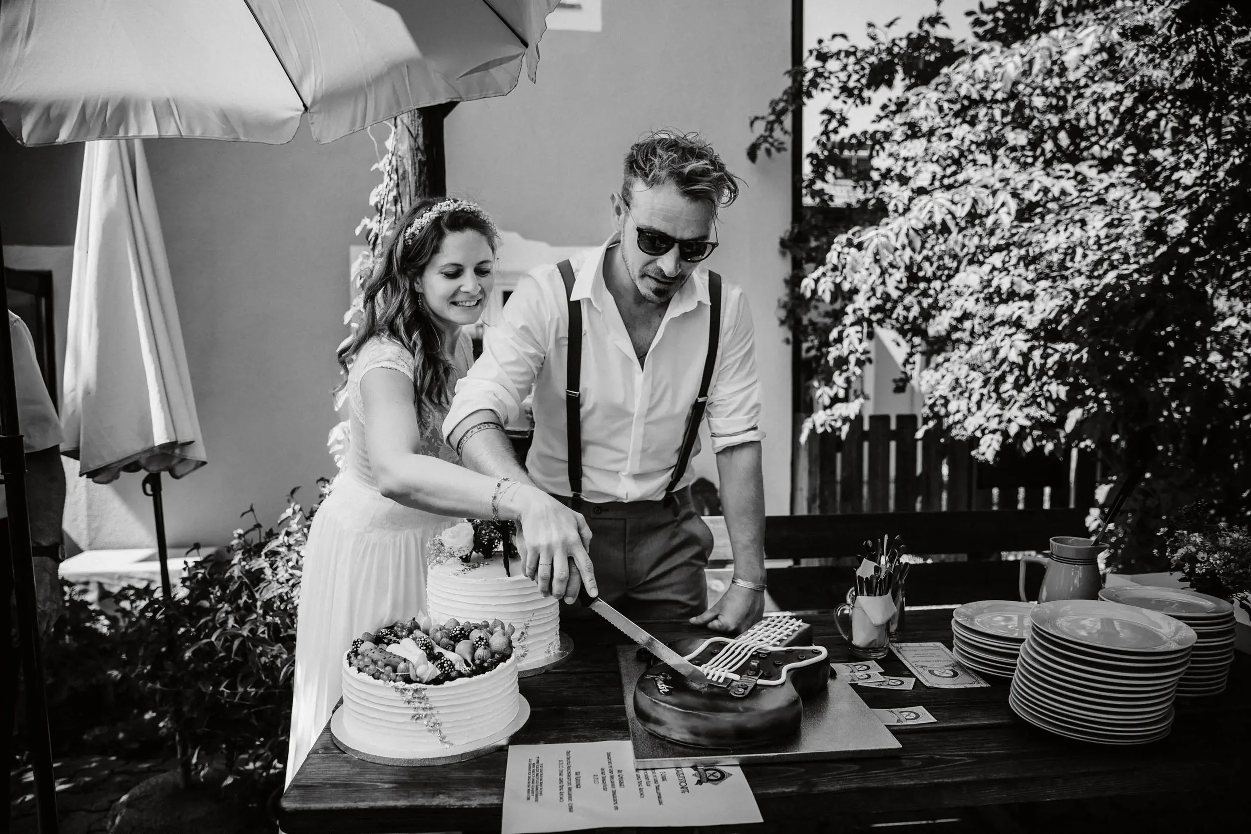Zwei Menschen, eine Frau und ein Mann, schneiden auf einer Terrasse eine Hochzeitstorte. Die Frau trägt ein weißes Kleid und ein Blumenkleid, während der Mann Sonnenbrille trägt und ein Hemd mit Hosenträgern. Es gibt einen Tisch mit weiteren Kuchen, 