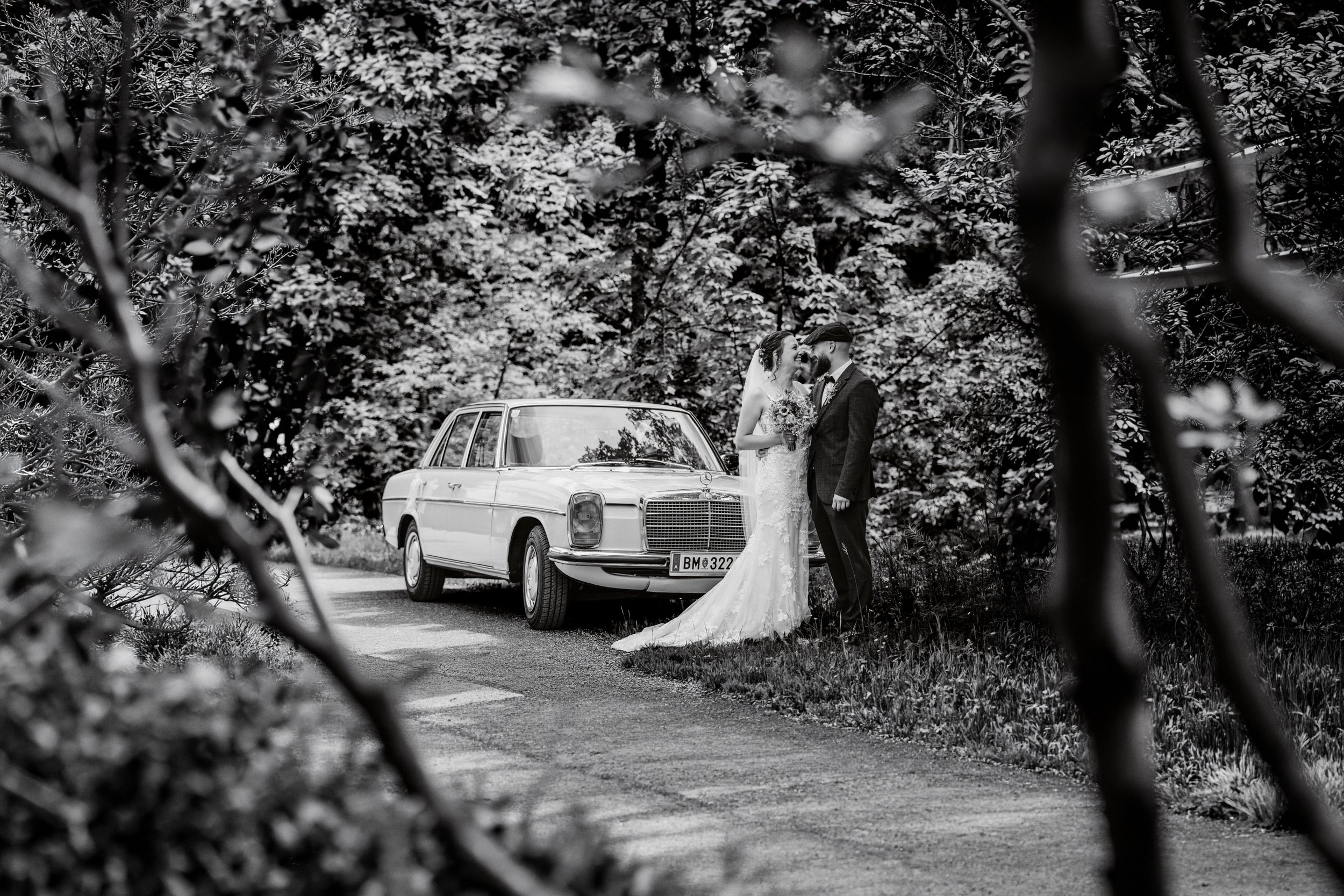 Ein Brautpaar während einer Hochzeit, stehend vor einem alten Mercedes-Benz Auto, umgeben von Bäumen, sichtbar durch ein Scherengitter im Vordergrund, in schwarz-weiß fotografiert.