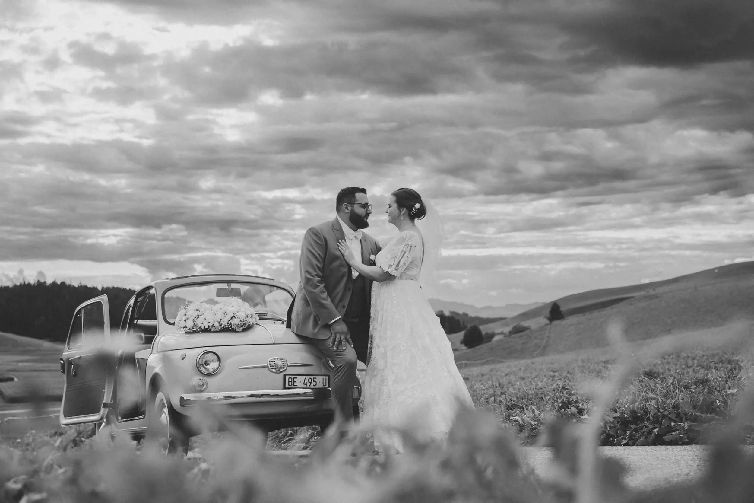 Ein glückliches Paar in Hochzeitskleidung steht vor einem alten Fiat-Auto auf einer ländlichen Straße, im Hintergrund bewölkter Himmel und weitläufige Felder.