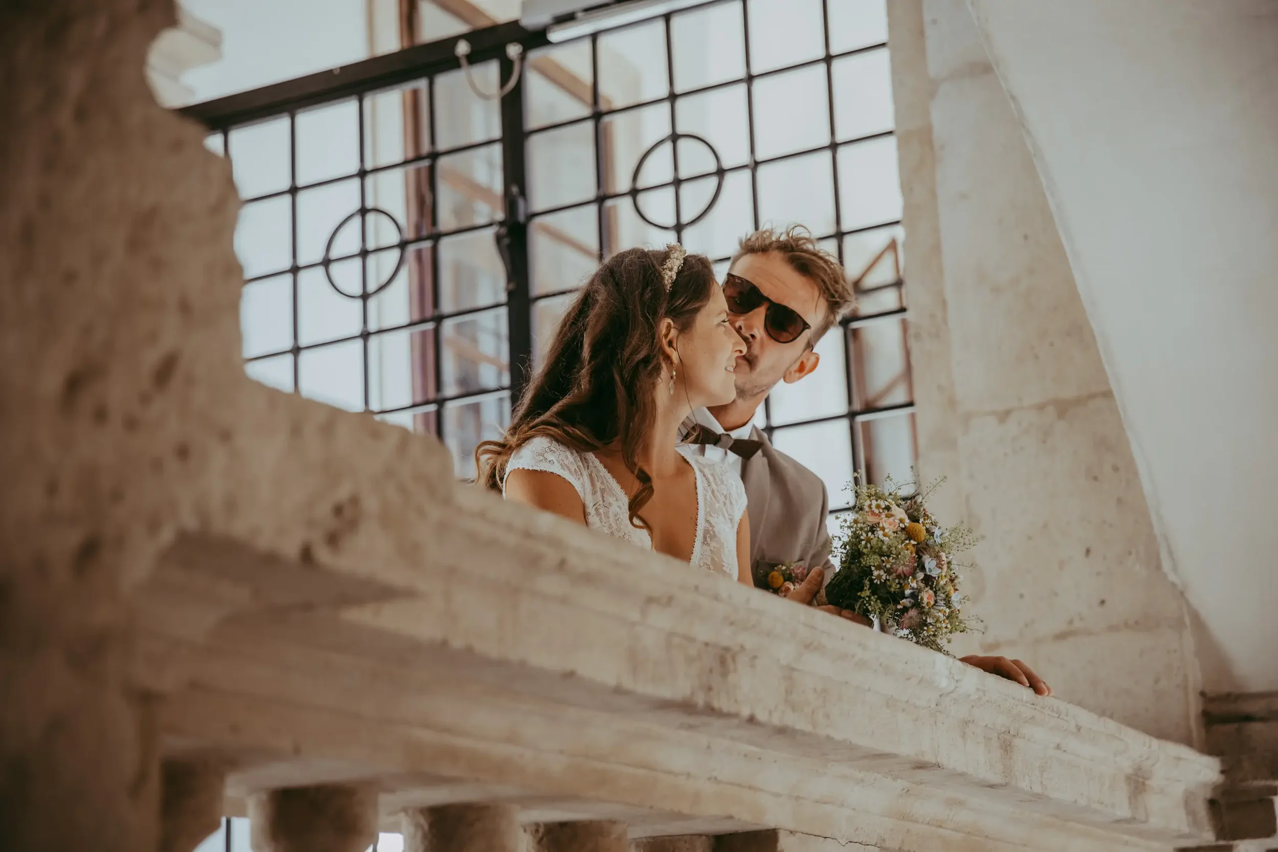 Ein Brautpaar während einer Hochzeit, das im Hintergrund durch ein großes Fenster mit Gitter blickt.