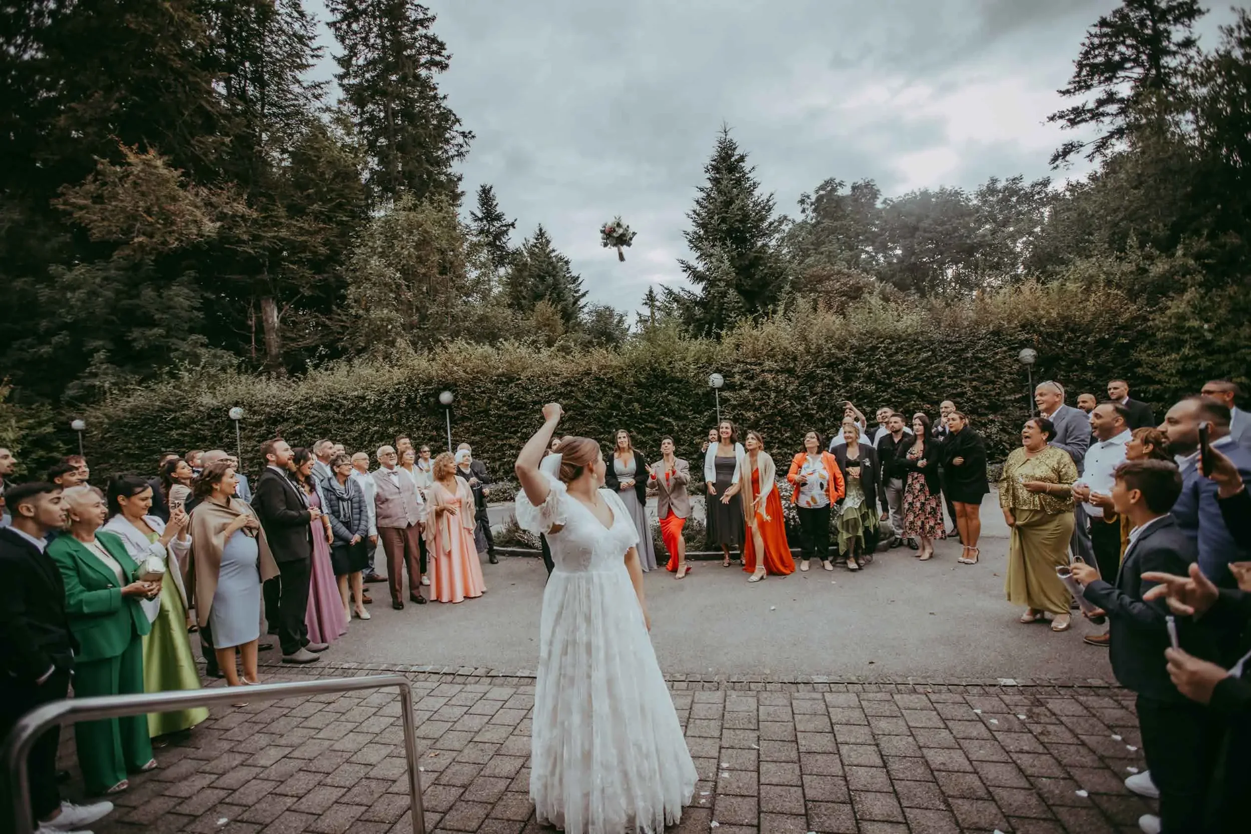Eine Gruppe von Menschen bei einer Hochzeitsfeier im Freien, im Hintergrund Bäume und ein Himmel mit Wolken, eine Frau in einem weißen Kleid steht im Mittelpunkt, während andere Menschen um sie herum stehen und Fotos machen.