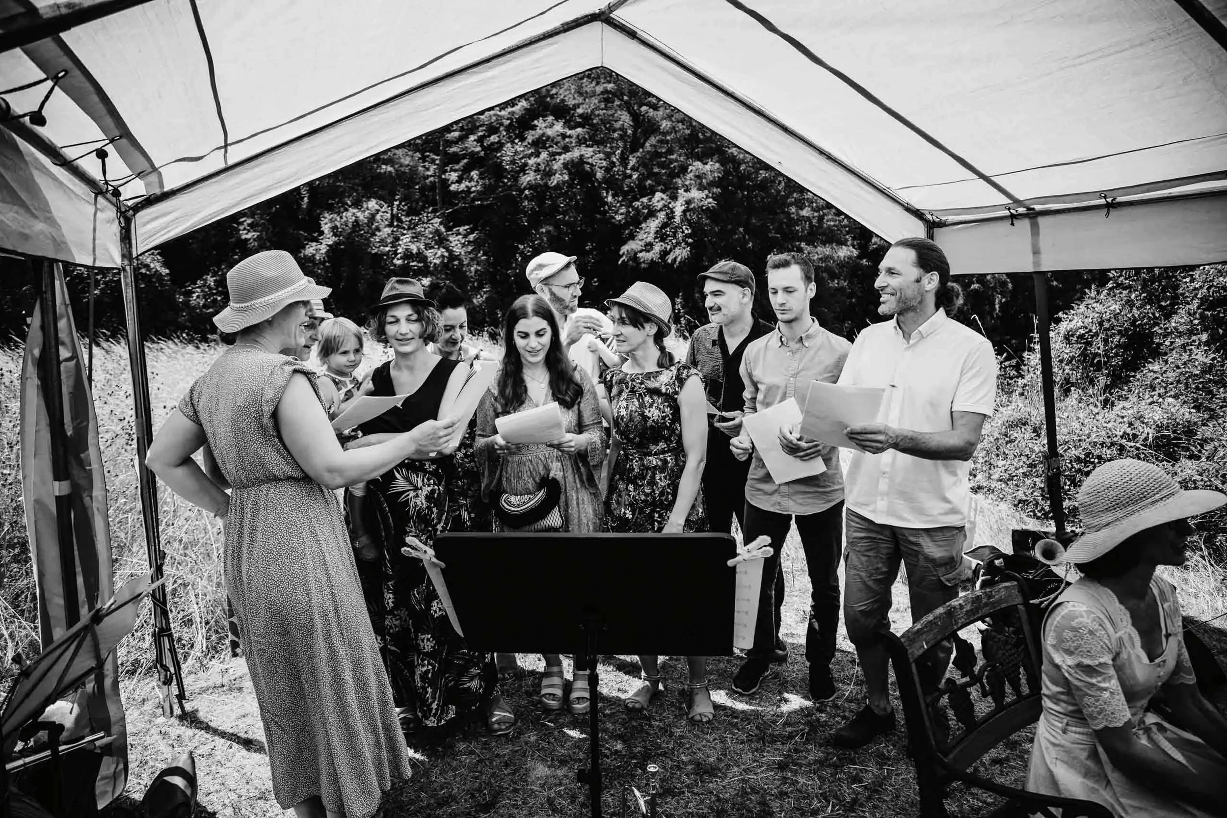 Eine Gruppe von Menschen beim Gesang oder Singen im Freien unter einem Zeltdach. Es sind Männer, Frauen und ein Kind, die Lieder oder Texte lesen oder singen, während sie um einen Notenständer versammelt sind.