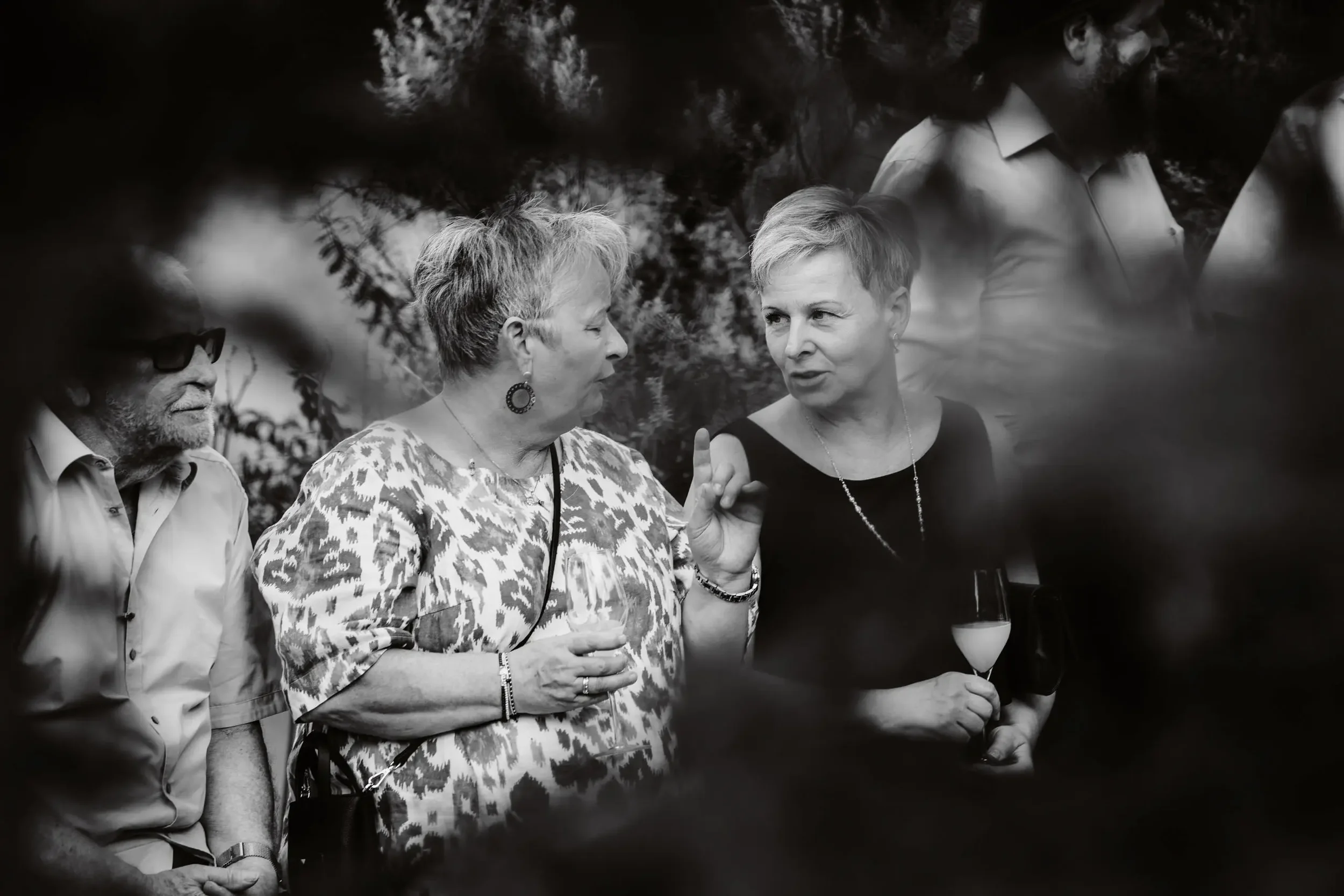 Fünf Menschen im Gespräch, drei Frauen und zwei Männer, beim Treffen im Freien. Zwei Frauen im Vordergrund, eine zeigt auf die andere. Die Szene ist in Schwarz-Weiß.