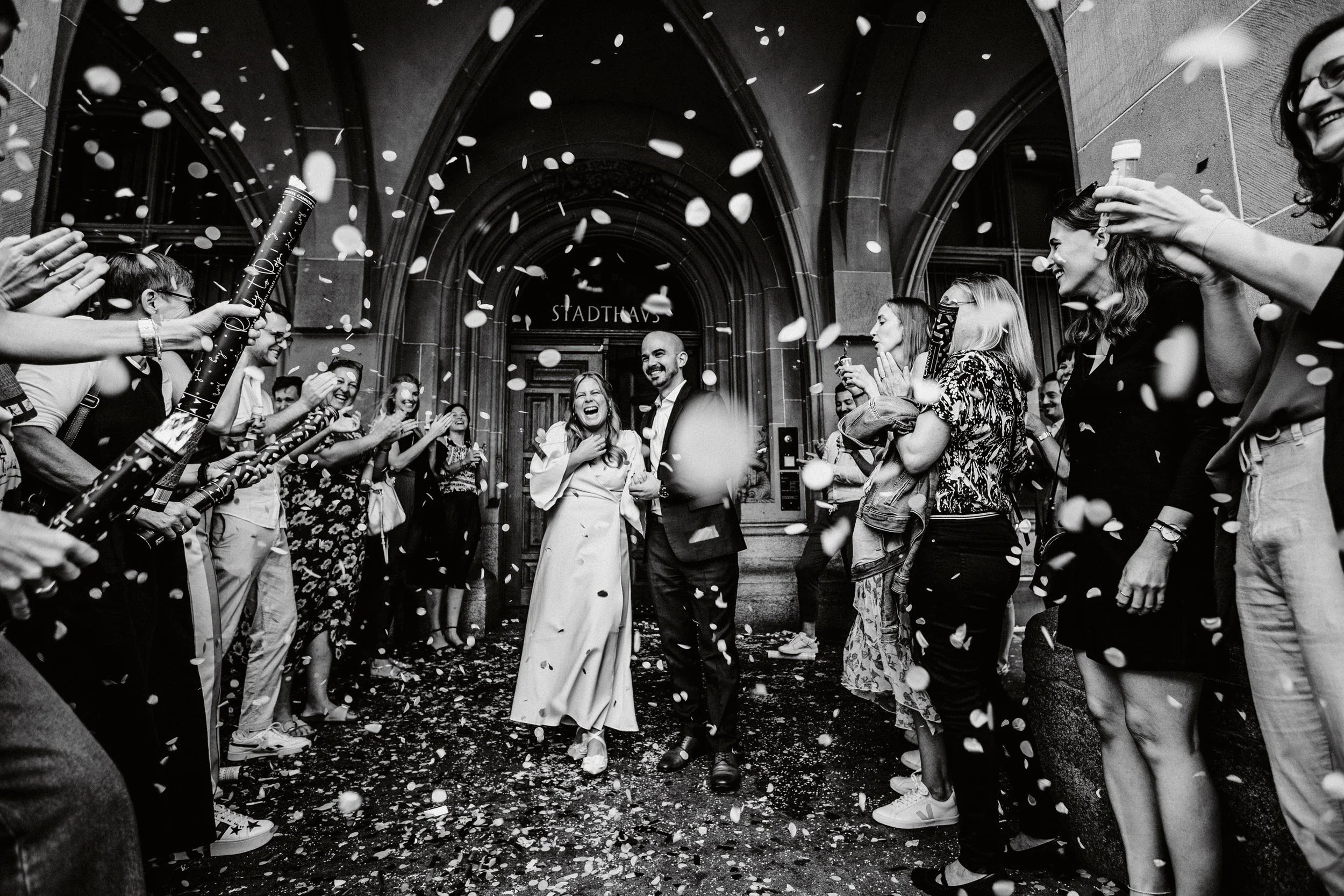 Feierliche Veranstaltung vor einem großen Gebäude mit Menschen, die alle lächeln, Konfetti werfen und sich umarmen, während es regnet, vermutlich bei einer Hochzeit oder einem Jubiläum.