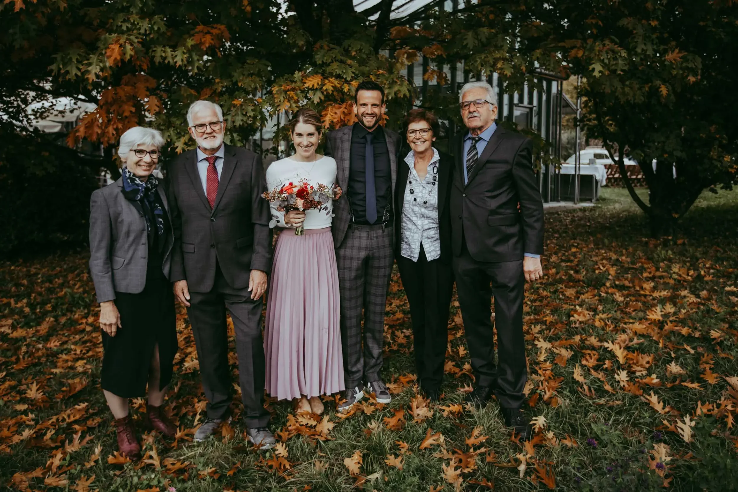 Eine Gruppe von sechs Menschen, darunter eine Frau mit einem Blumenstrauß, steht im Freien vor einem Baum mit herbstlichen Blättern und lächelt in die Kamera.