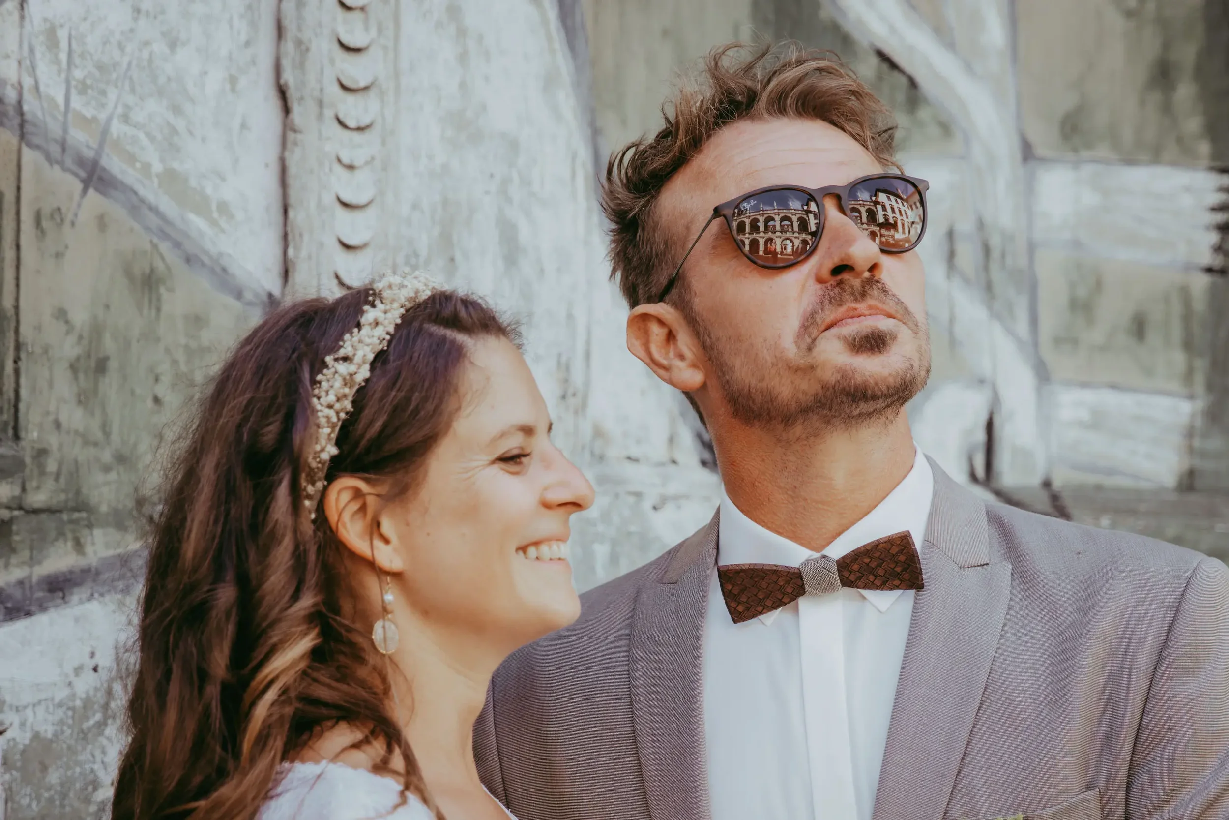Ein Mann in einem grauen Anzug mit Fliege und Sonnenbrille steht neben einer lächelnden Frau mit langen, lockigen Haaren und einem Haarschmuck vor einer alten Mauer.