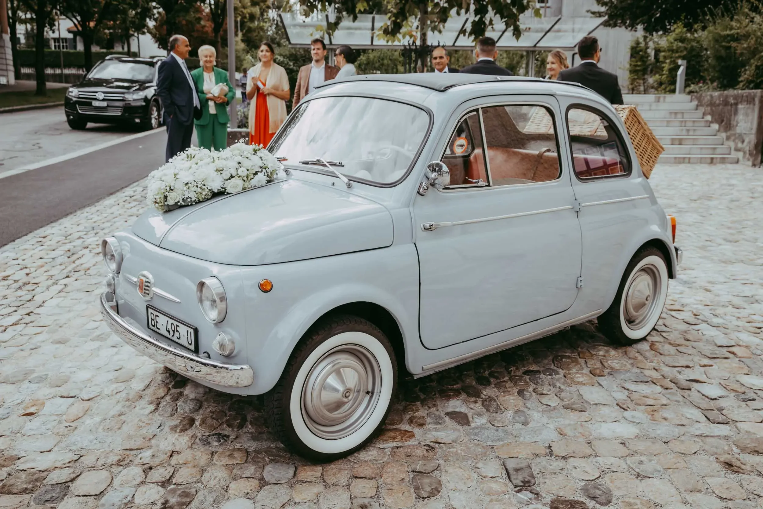 Ein weißer Oldtimer mit Blumen auf der Motorhaube ist auf einer gepflasterten Straße geparkt. Im Hintergrund stehen Menschen in formeller Kleidung und unterhalten sich. Es sieht nach einer festlichen Veranstaltung oder Hochzeit aus.