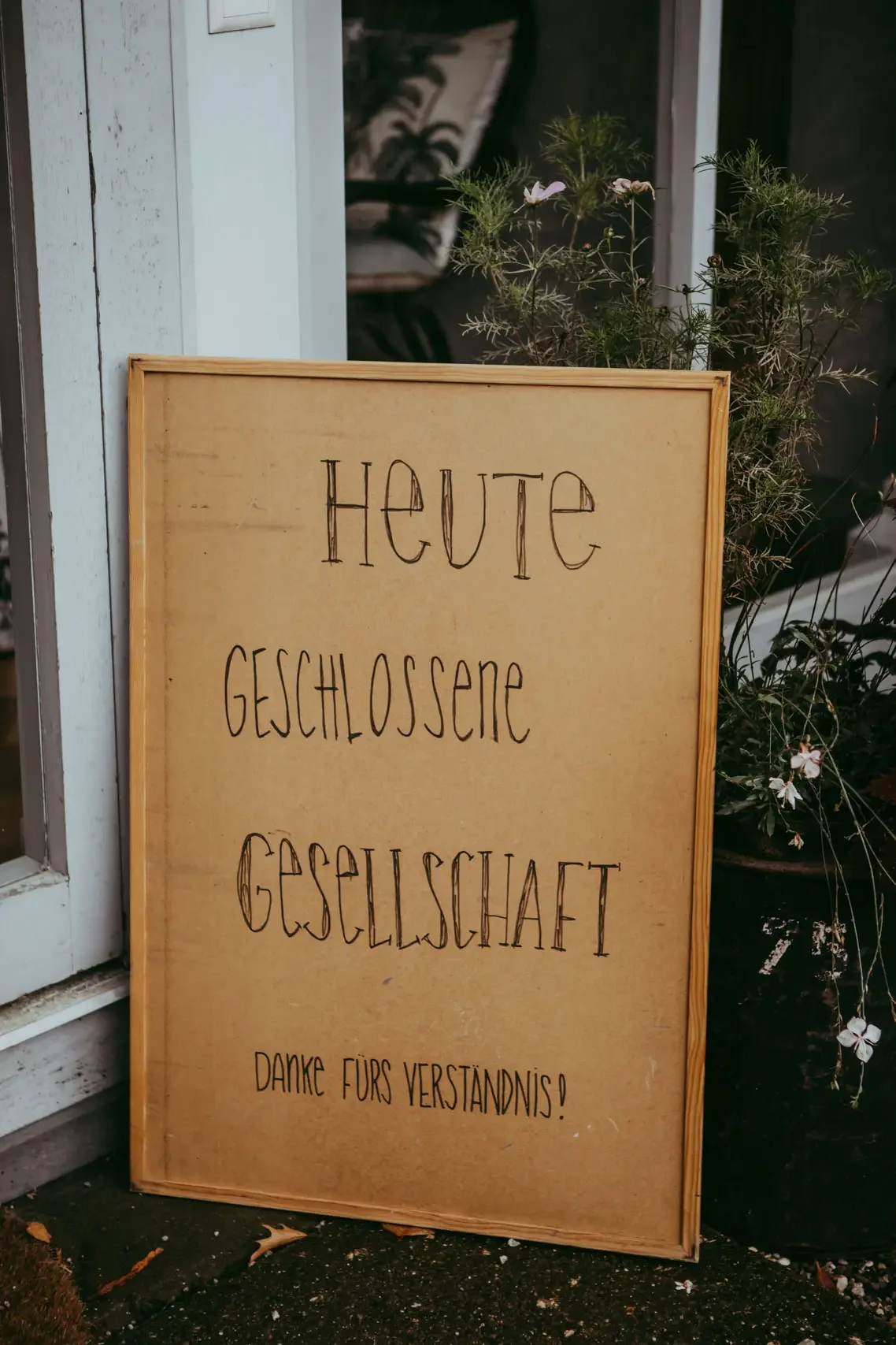 Ein Holzschild mit der Aufschrift 'Heute geschlossen, Gesellschaft, Danke fürs Verständnis!' steht vor einem Gebäude, umgeben von Pflanzen.