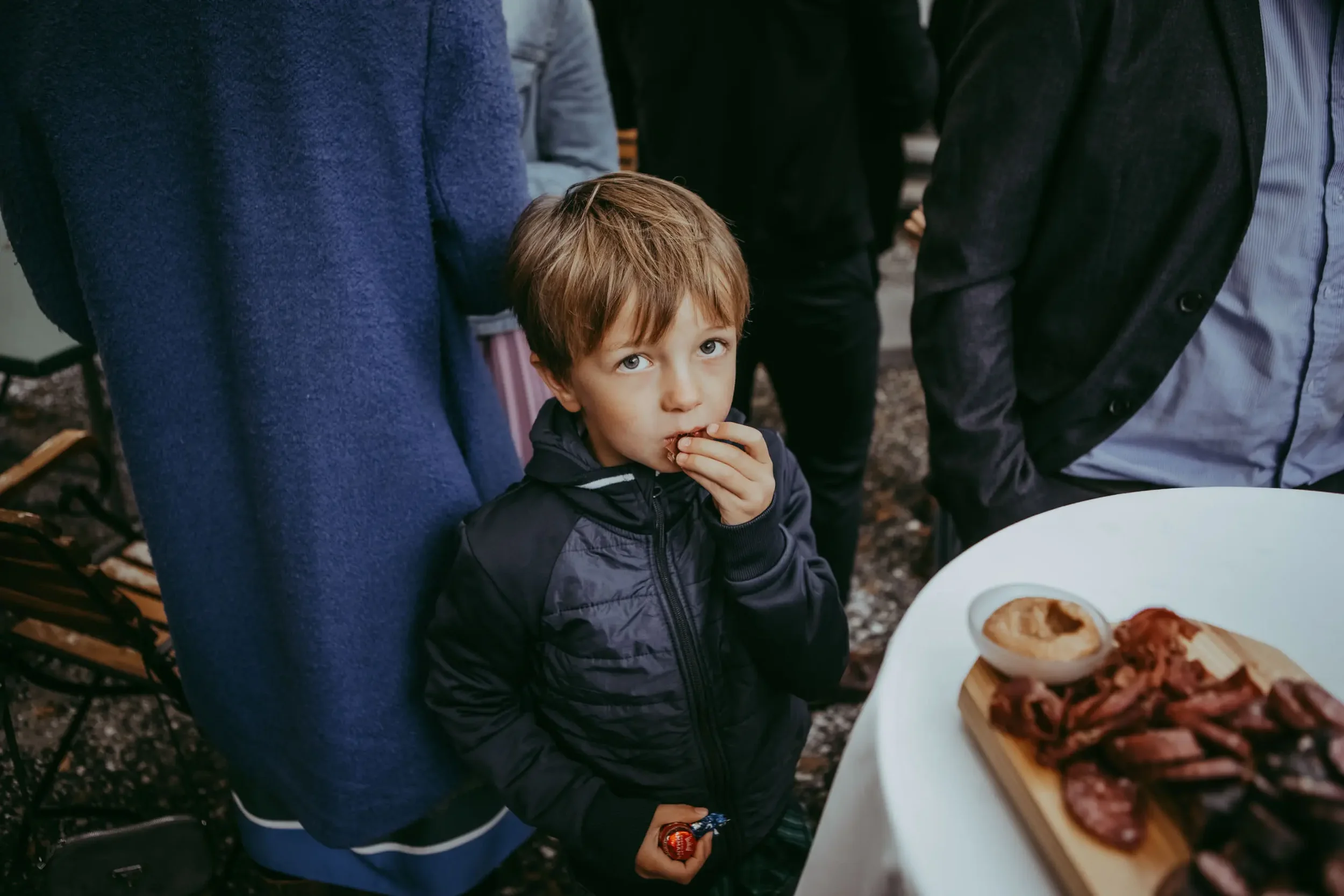 Ein Junge isst ein Stück Pizza bei einer Zusammenkunft im Freien, umgeben von Erwachsenen, mit einer Pizza auf dem Tisch im Vordergrund.