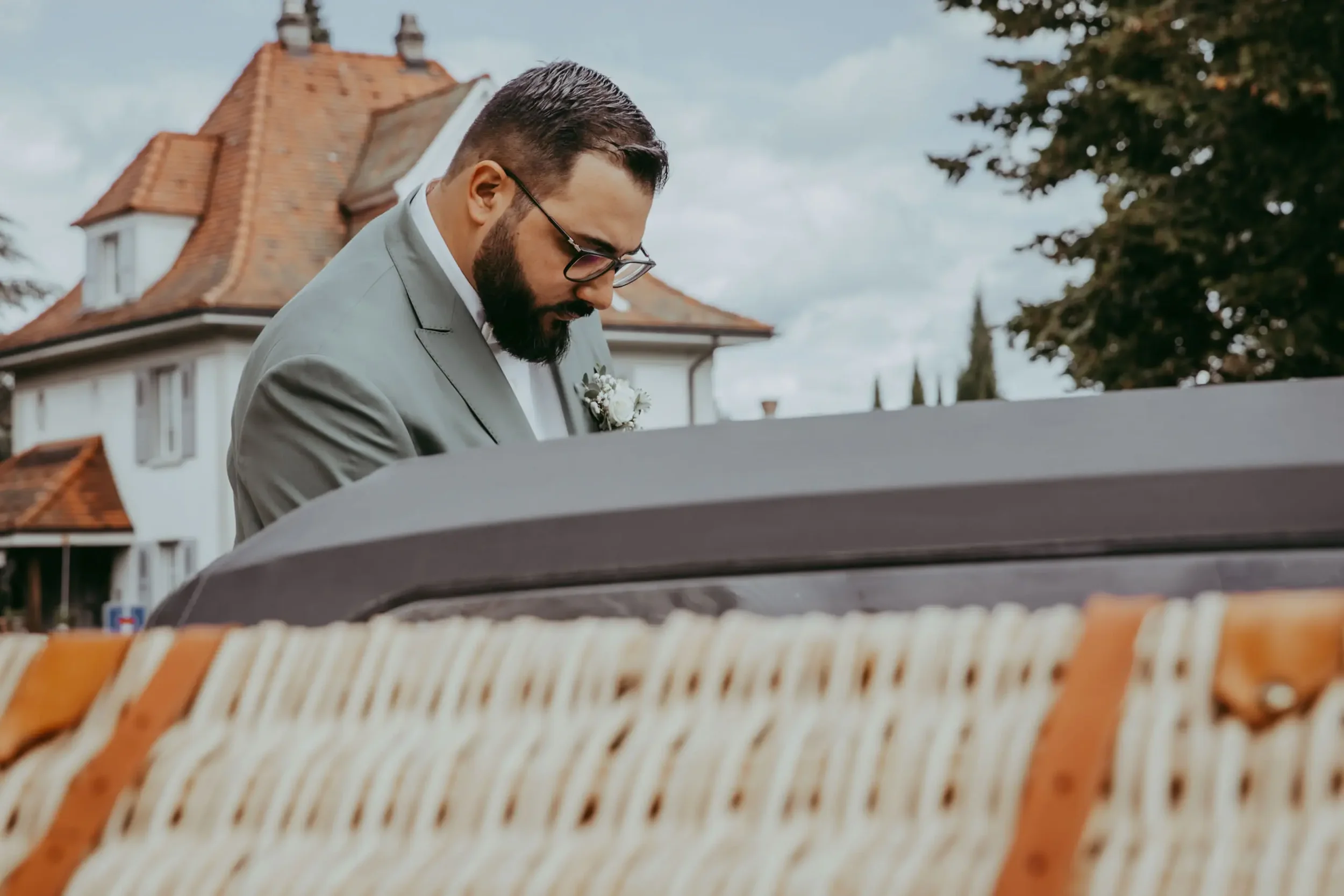 Ein Mann mit Brille und Bart, trägt einen Anzug mit weißem Hemd, spielt Klavier im Freien vor einem historischen Gebäude mit rotziegeligem Dach und weißen Wänden, umgeben von Bäumen.