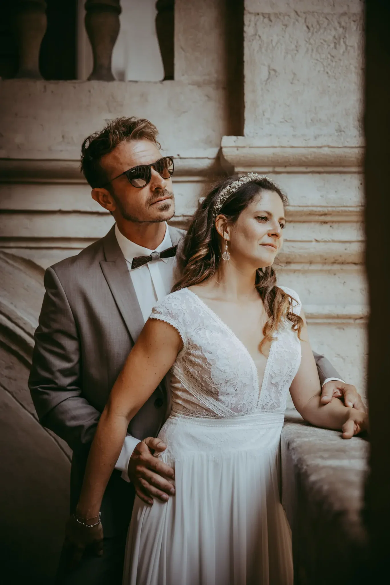 Ein Mann in einem Anzug mit Sonnenbrille hält eine Frau in einem weißen Brautkleid fest, beide stehen an einer Steintreppe bei einem Hochzeitsereignis.
