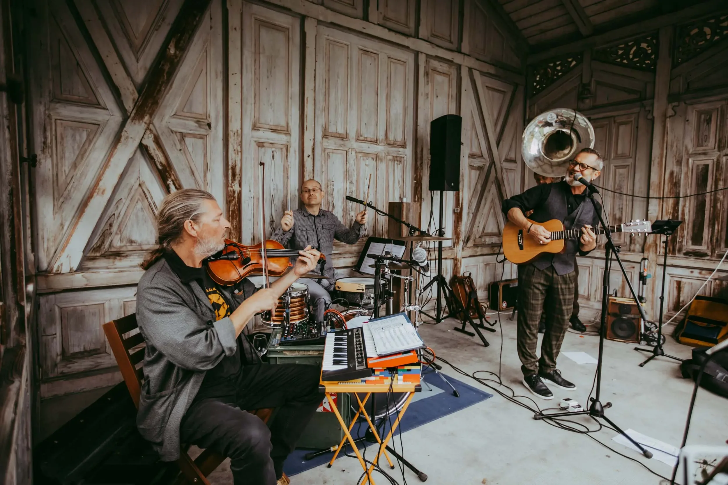Drei Musiker spielen in einem Raum mit Holzwänden. Ein Musiker spielt Geige, ein Musiker sitzt hinter einem elektronischen Musikgerät, ein dritter Musiker singt und spielt Gitarre.