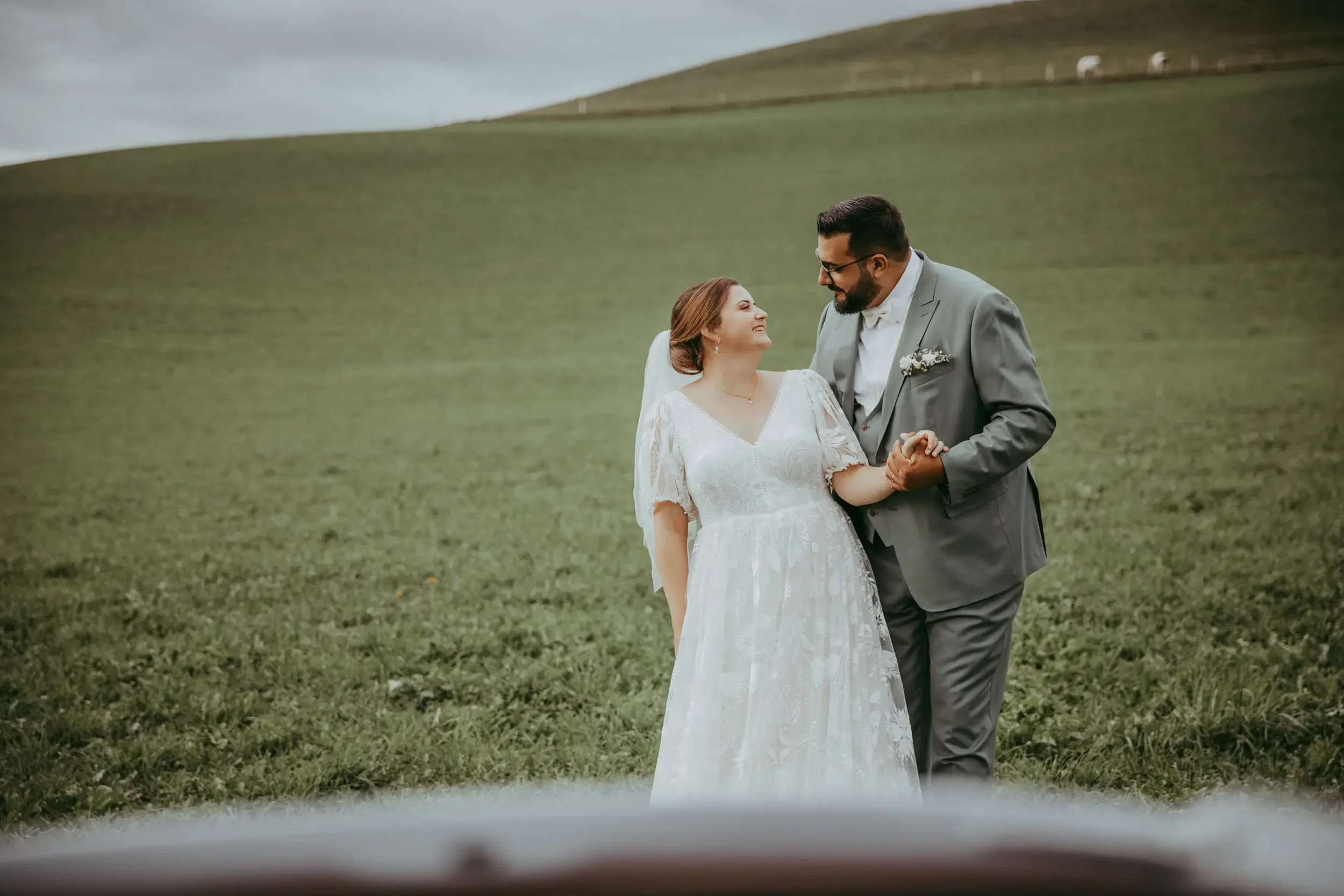 Ein glückliches Brautpaar hält Händchen auf einer grünen Wiese mit Hügeln im Hintergrund, bewölktem Himmel, bei einer Hochzeit im Freien.